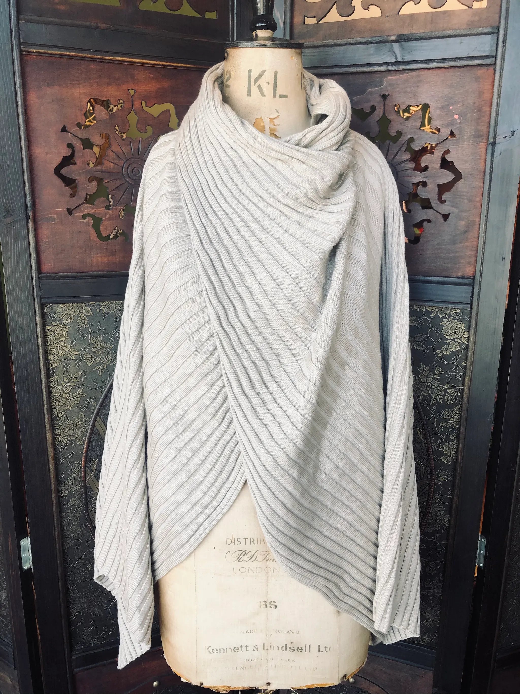 Broochella, COS, One Size,Wool ribbed knit, Beige poncho. Wrap-front design,
Sculptural texture, in excellent pre-owned condition. It is pictured on a vintage mannequin. The background is an ornate, vintage, dark wood room divider. 