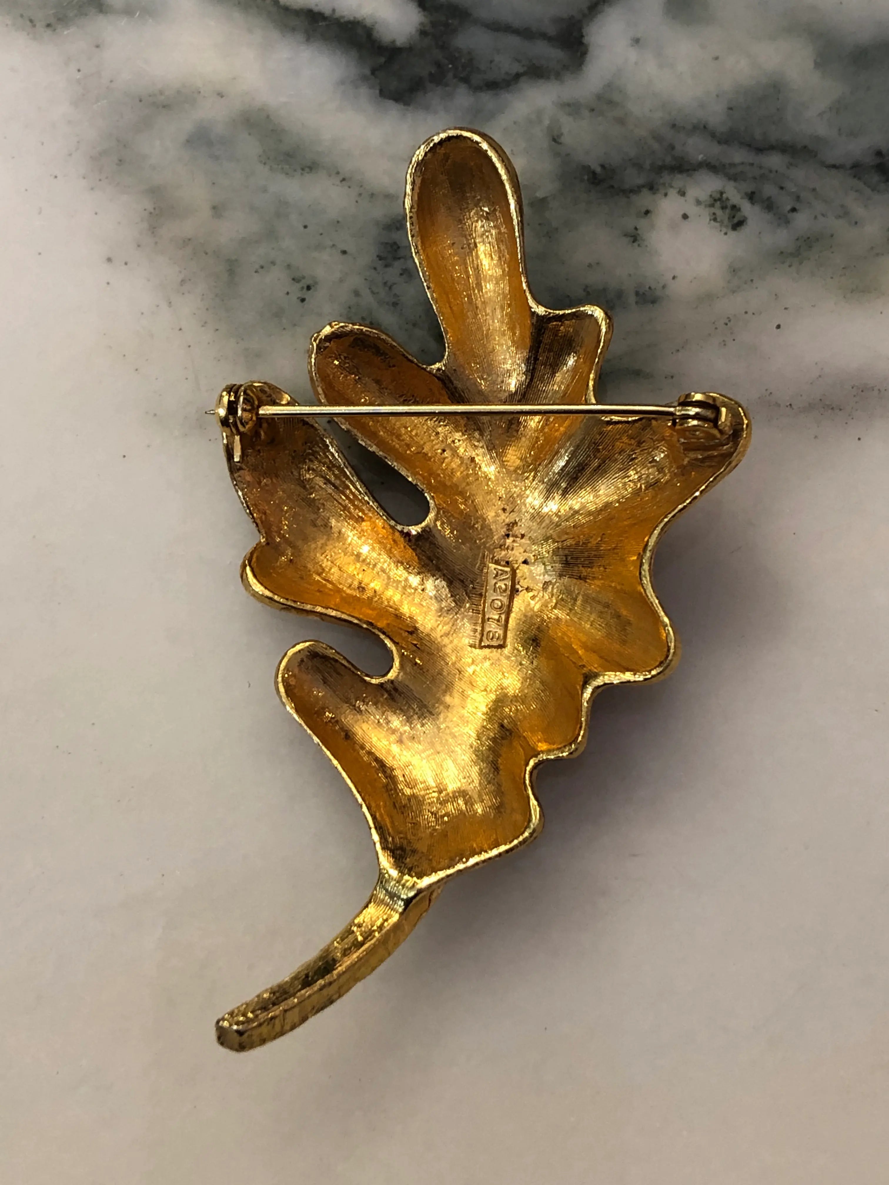 The back view of Broochella Cosima Enamel Leaf Brooch featuring ruby-red and navy enamel, gold-tone detailing and a crystal-set stem on a cream and grey marble background.