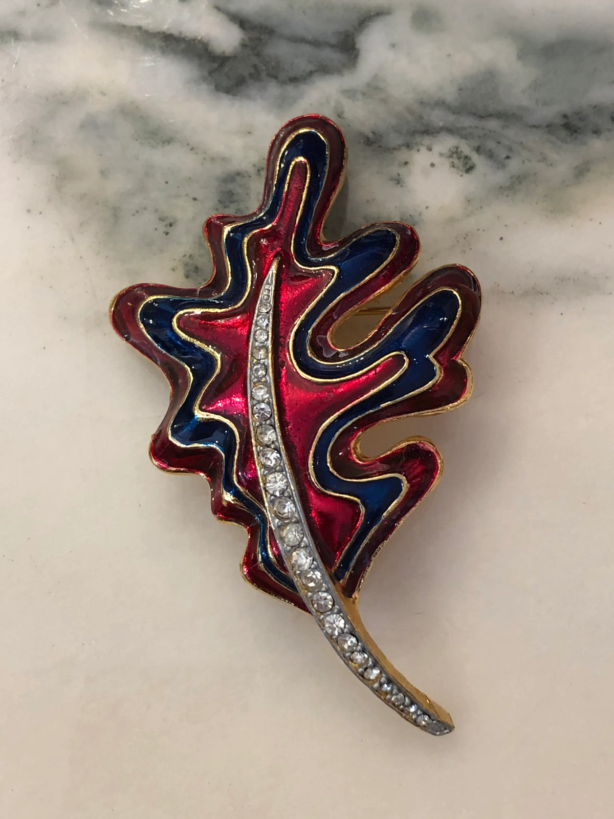 Broochella Cosima Enamel Leaf Brooch featuring ruby-red and navy enamel, gold-tone detailing and a crystal-set stem on a cream and grey marble background.