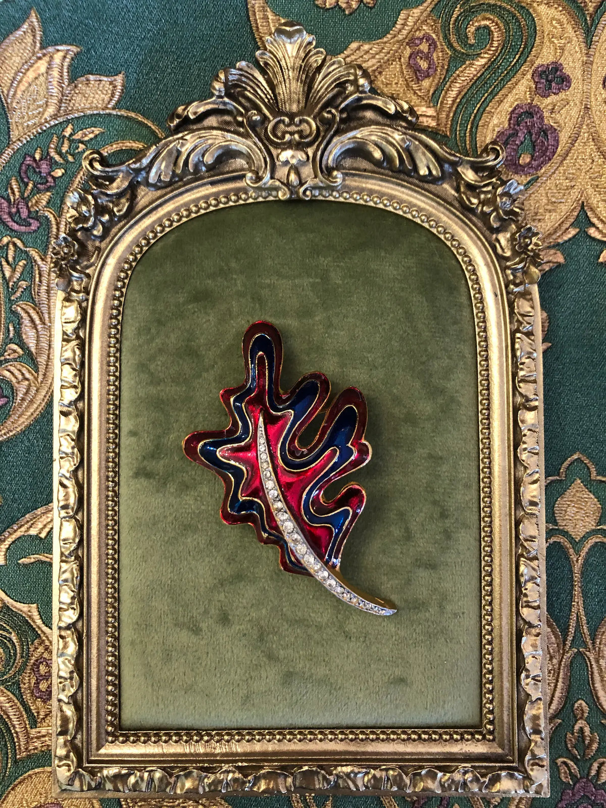 Broochella Cosima Enamel Leaf Brooch featuring ruby-red and navy enamel, gold-tone detailing and a crystal-set stem. It is pictured on a green velvet tray with an ornate gold frame. The background is green and gold damask wallpaper