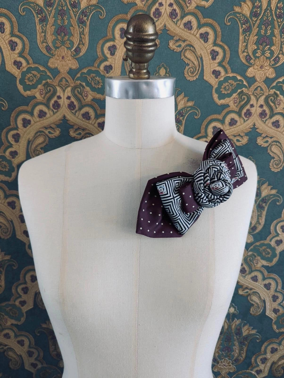 A Broochella Bolivar vintage upcycled necktie styled on a cream fabric dress form mannequin, featuring a burgundy and white geometric pattern with a central fabric rose embellishment. The background is green and gold damask wallpaper.