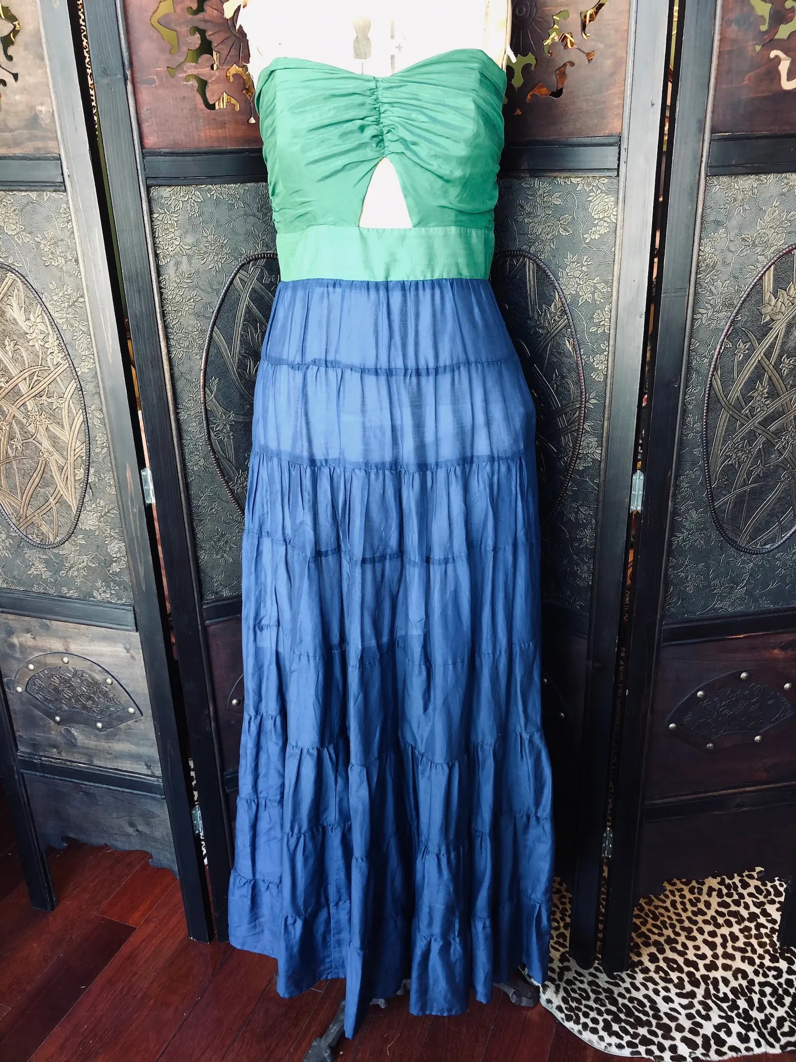 Broochella Brand: Cristina – Beautiful Life
Strapless maxi dress.Green ruched bodice. Blue tiered maxi skirt. Centre cut-out detail. 70% cotton, 30% silk
Lightweight, breathable fabric
Size: Small. It is presented on a vintage mannequin. The background is a decorative wooden room divider.