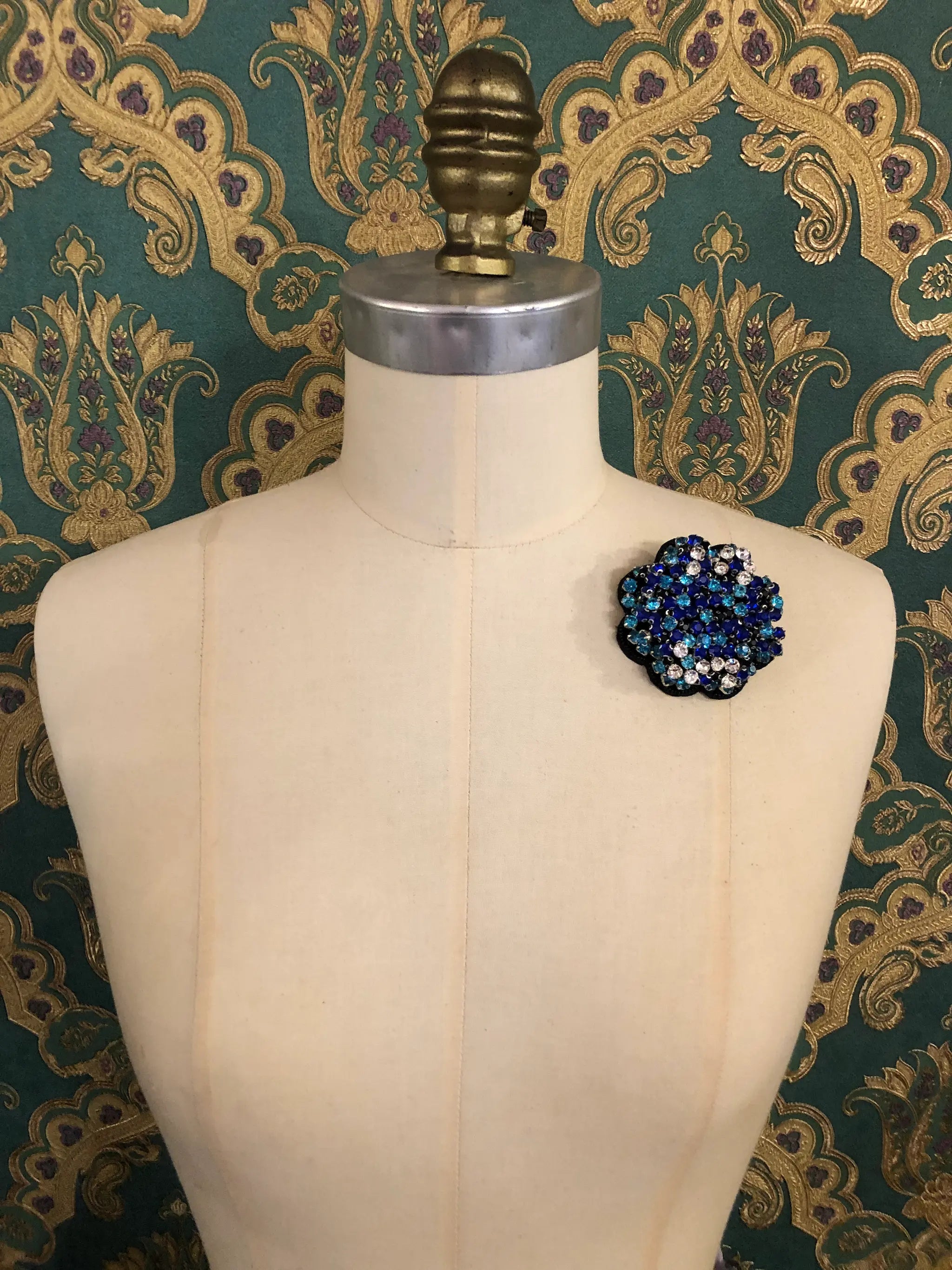 A Broochella Crystal Blooms small floral brooch with dark blue, sky blue, and white rhinestones, displayed on a cream-colored dress form against a dark green and gold patterned wallpaper background.