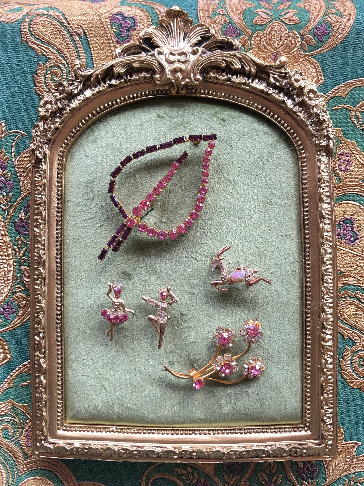 Broochella Da Vinci Quint Vintage Brooch Set featuring five pink and purple crystal brooches displayed on sage green velvet in an ornate vintage gold frame against a teal and gold patterned background.