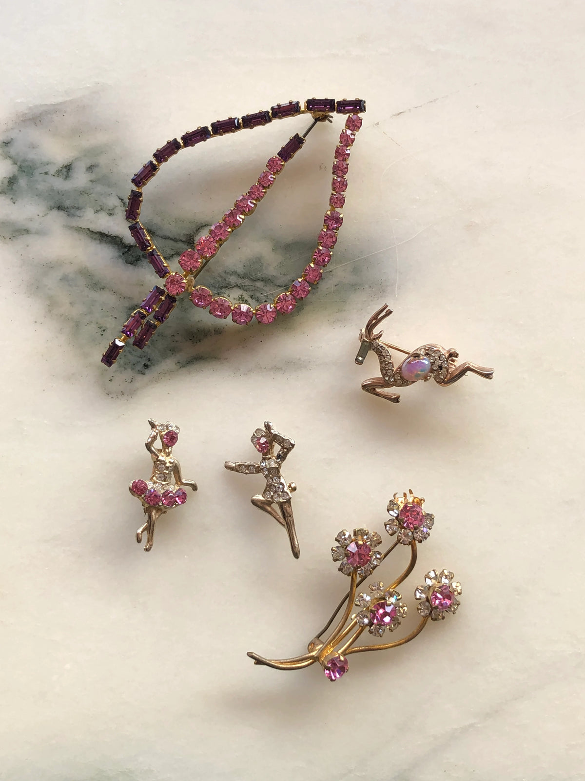 Broochella Da Vinci Quint Vintage Brooch Set featuring five delicate pink and purple crystal designs including an initial, ballerinas, leaping deer and floral spray, photographed on a white marble background.