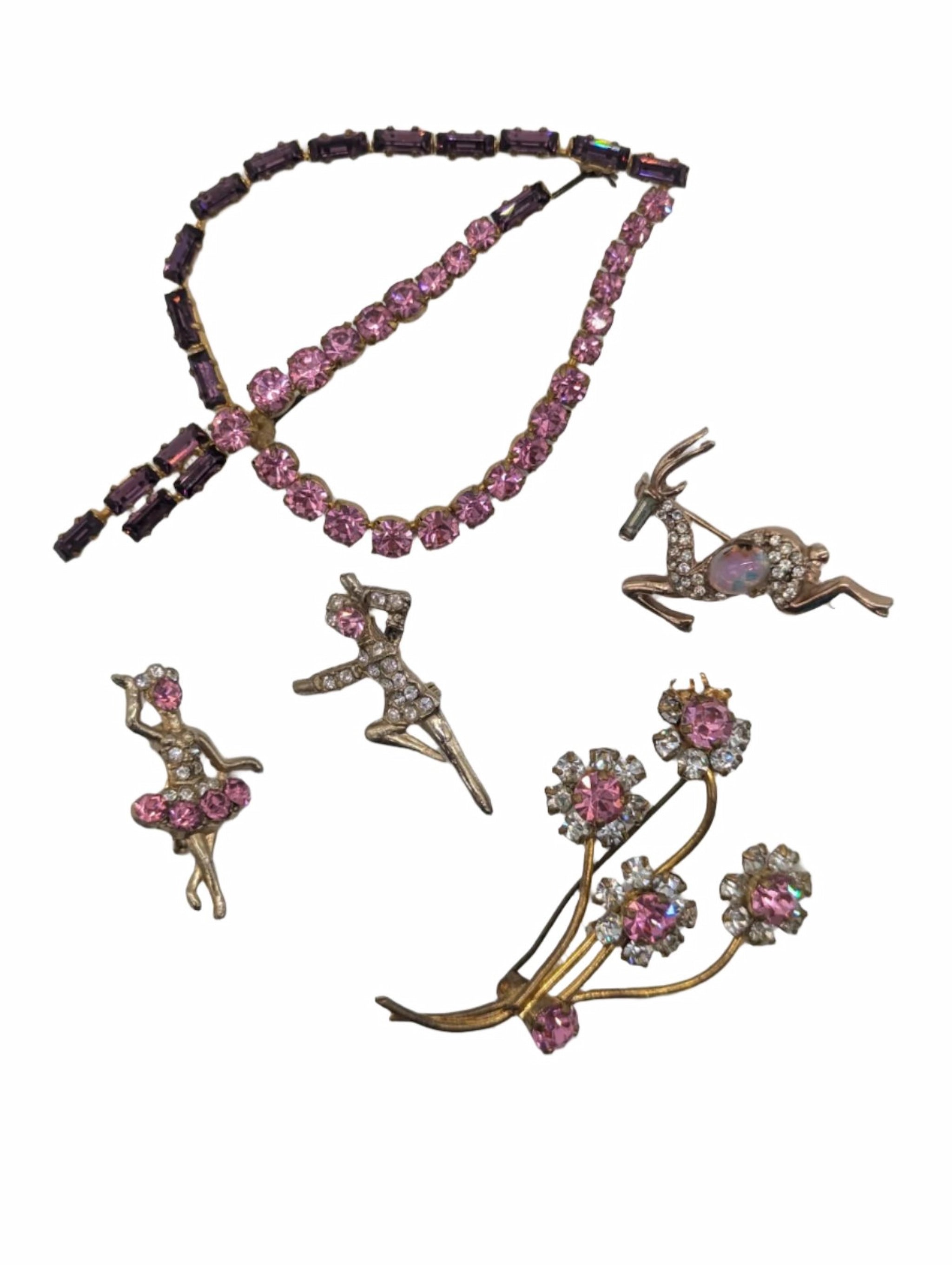 Broochella da vinci five piece vintage brooch set in gold tone metal, with pink crystals. The set includes flowers, dancing figures, a leaf and a deer. The background is white. 