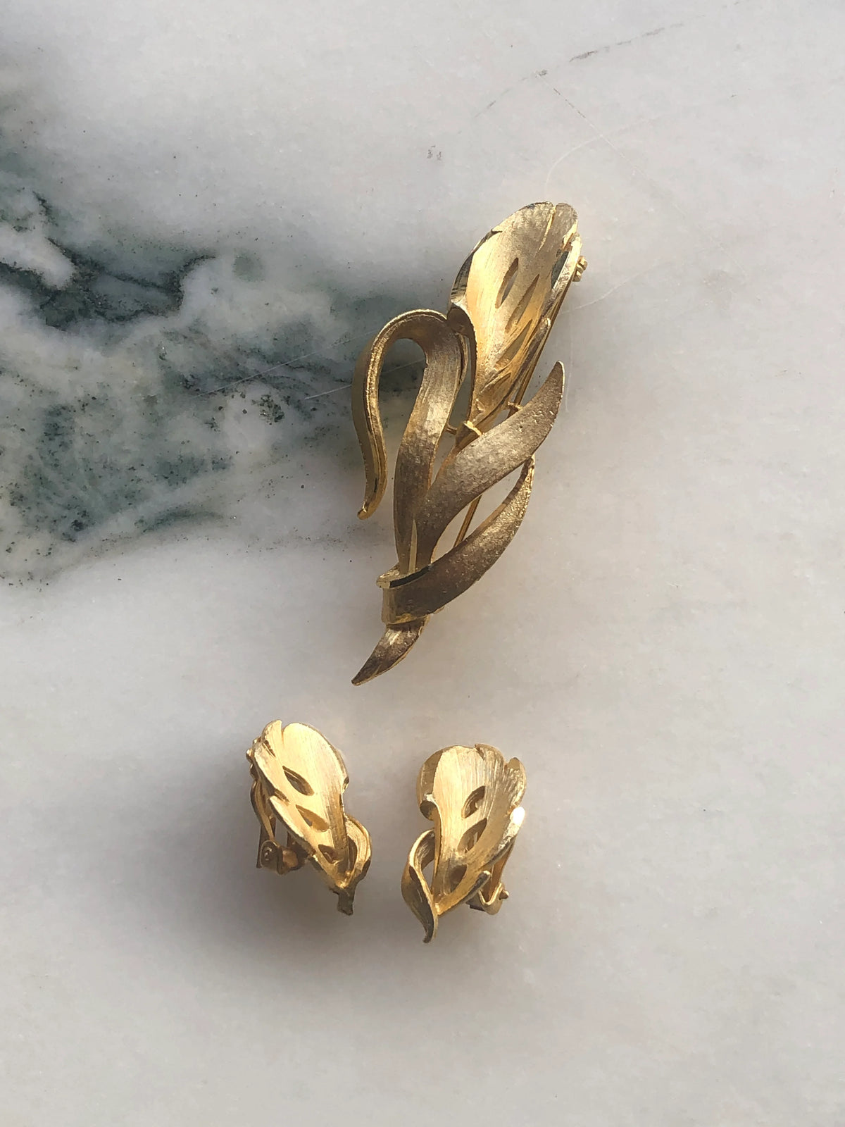 Broochella Da Vinci Vintage Brooch and Clip-on Earring Set featuring a large sculptural gold-tone brooch with two matching earrings, photographed on a white marble background with soft grey-green veining.