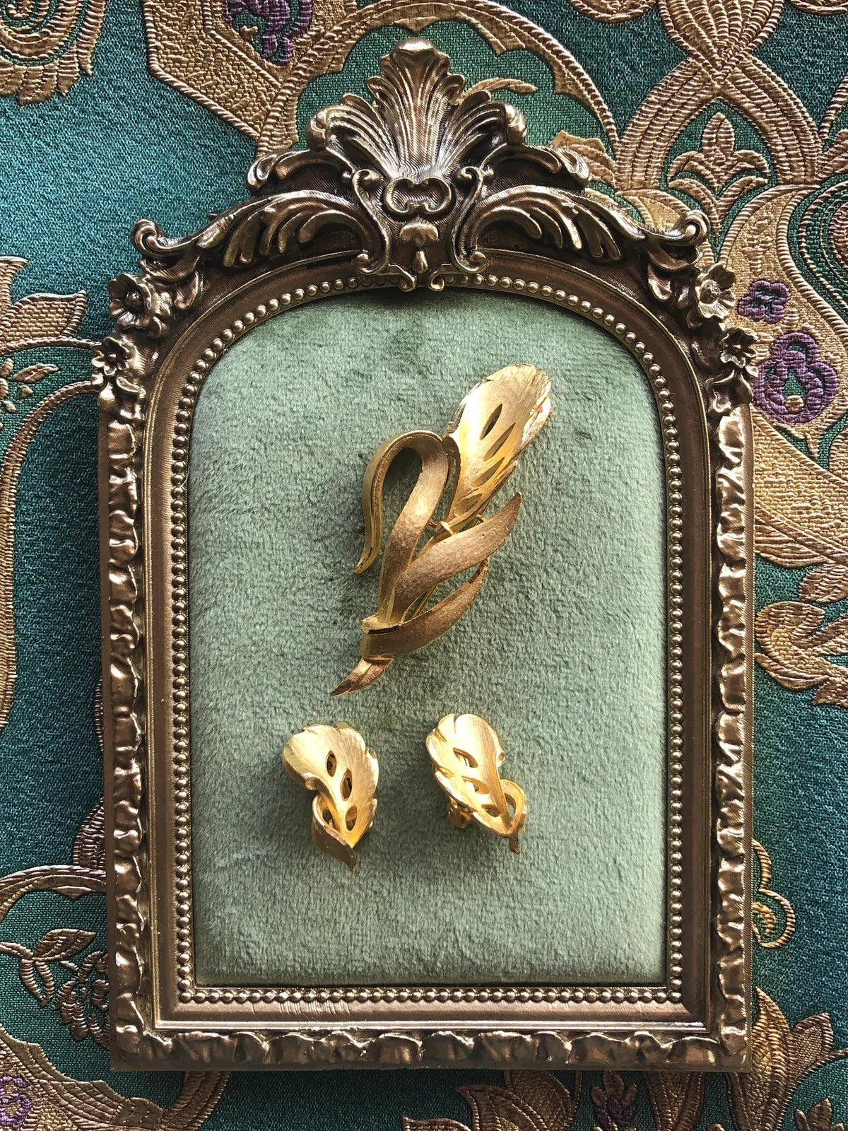 Broochella Da Vinci Vintage Brooch and Clip-on Earring Set in sculptural gold-tone metal with elegant leaf and ribbon forms, displayed on sage green velvet in an ornate antique gold frame against green and gold patterned fabric.
