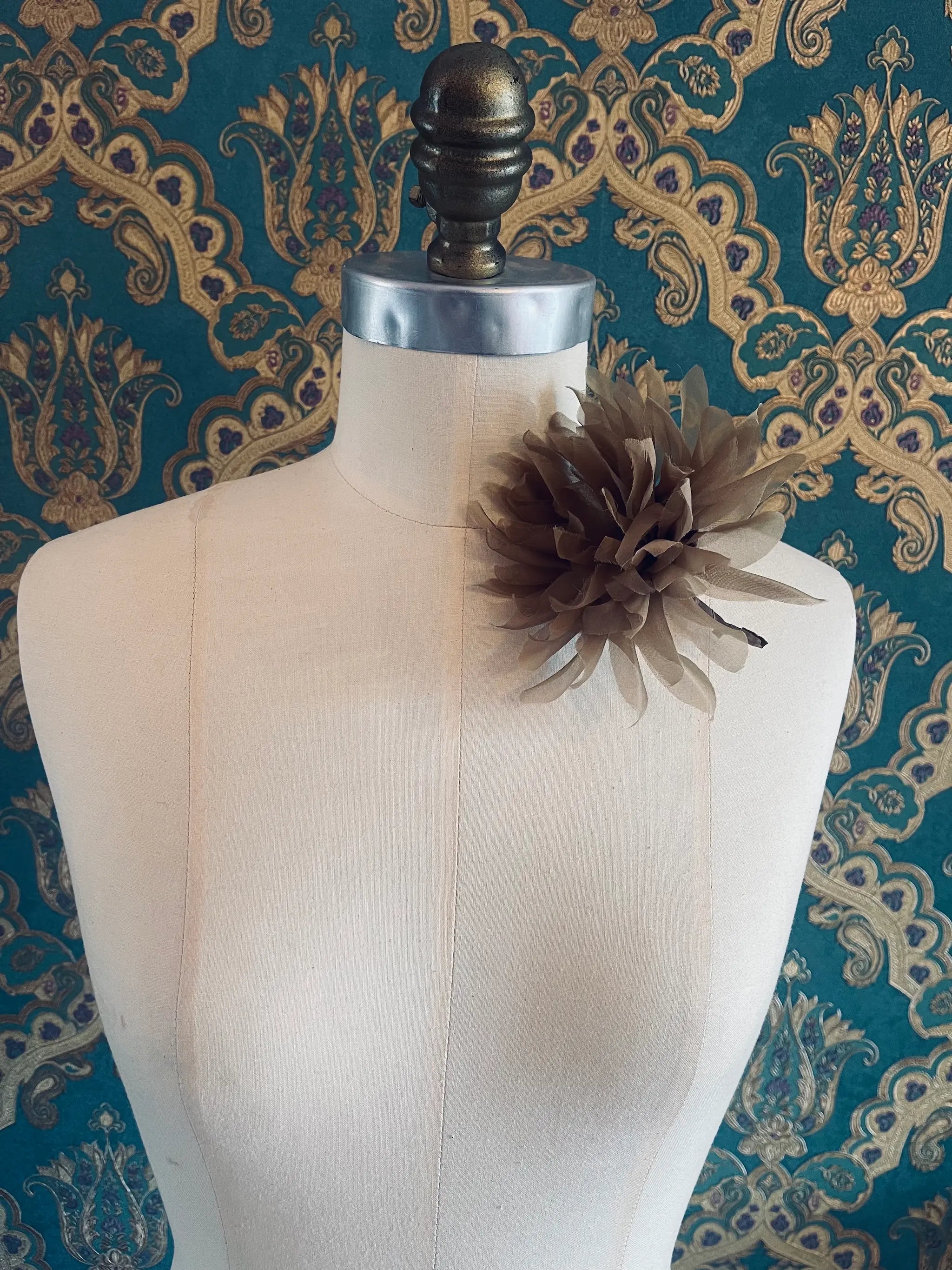Broochella Dahlia handmade fabric flower brooch in brown. Styled on a mannequin. The background is green and gold damask wallpaper