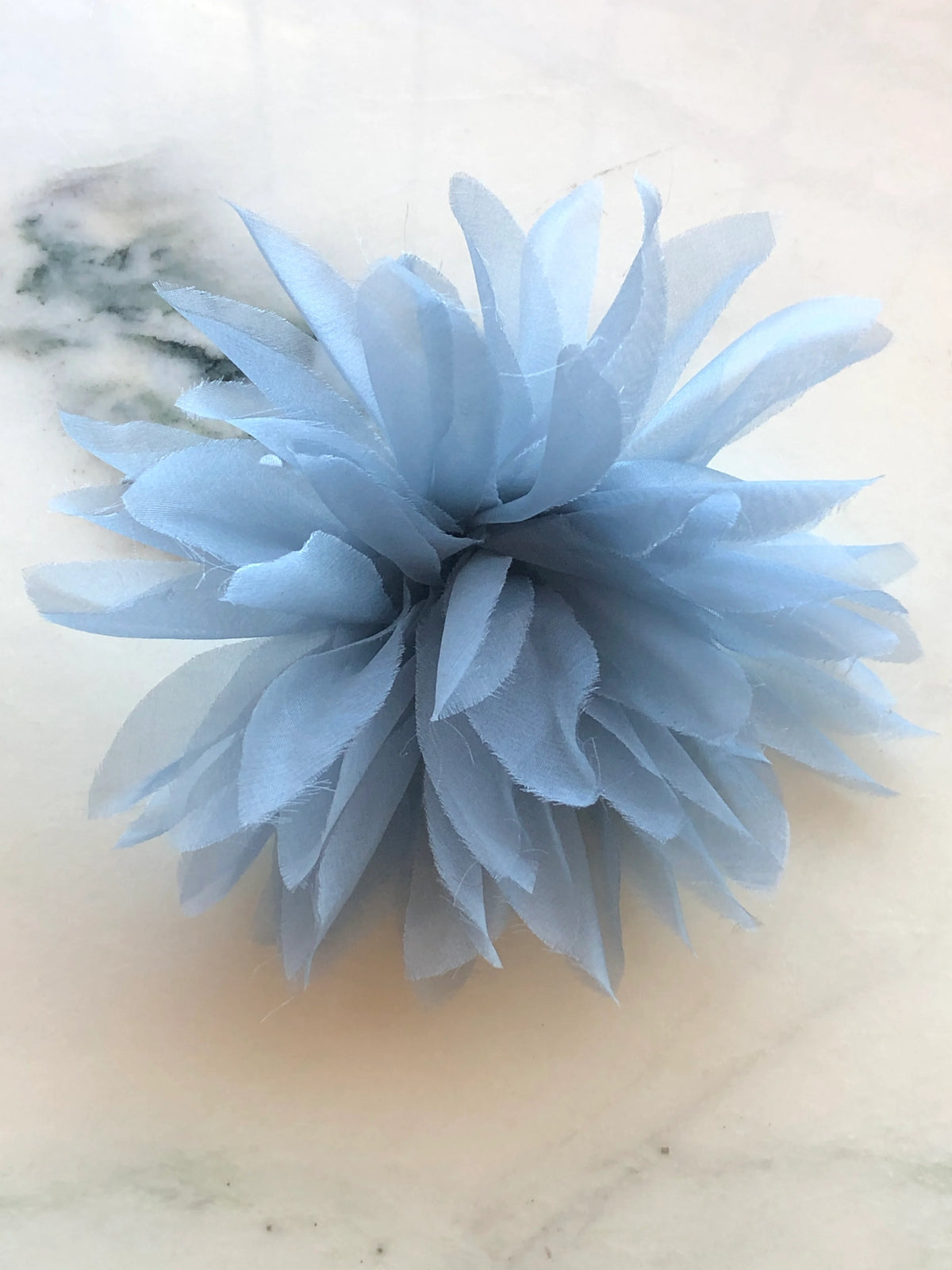 Broochella Dahlia Flower Brooch Pin in soft powder blue, featuring full layers of long pointed fabric petals with softly frayed edges on a cream marble background.