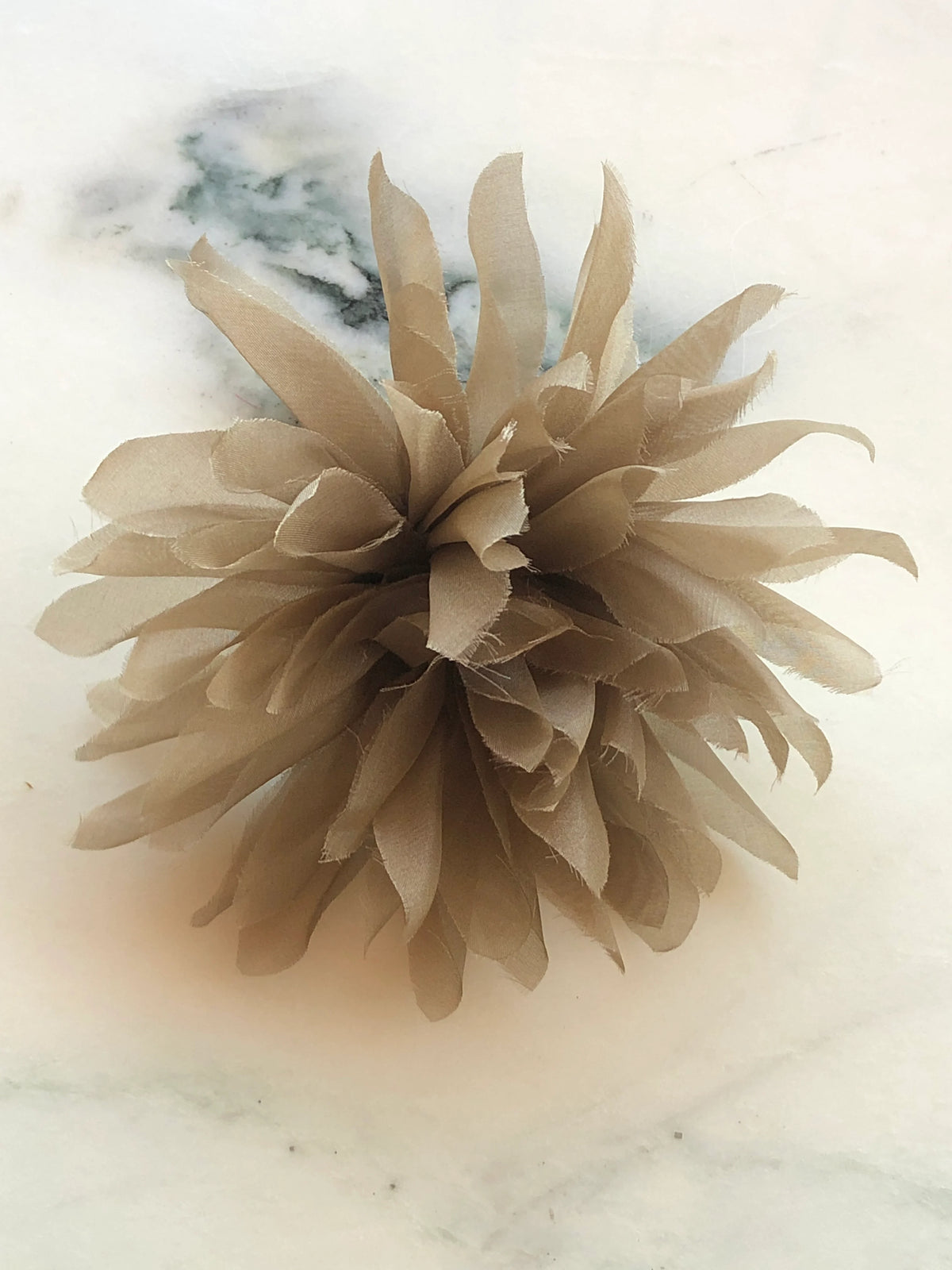 Broochella Dahlia Flower Brooch Pin in soft taupe brown, featuring full layers of long pointed fabric petals with softly frayed edges on a cream marble background.
