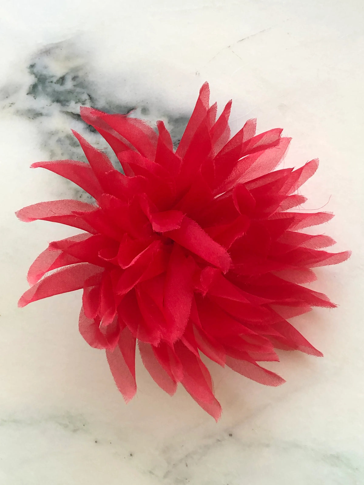Broochella Dahlia Flower Brooch Pin in vibrant red, featuring full layers of long pointed fabric petals with softly frayed edges on a cream marble background.