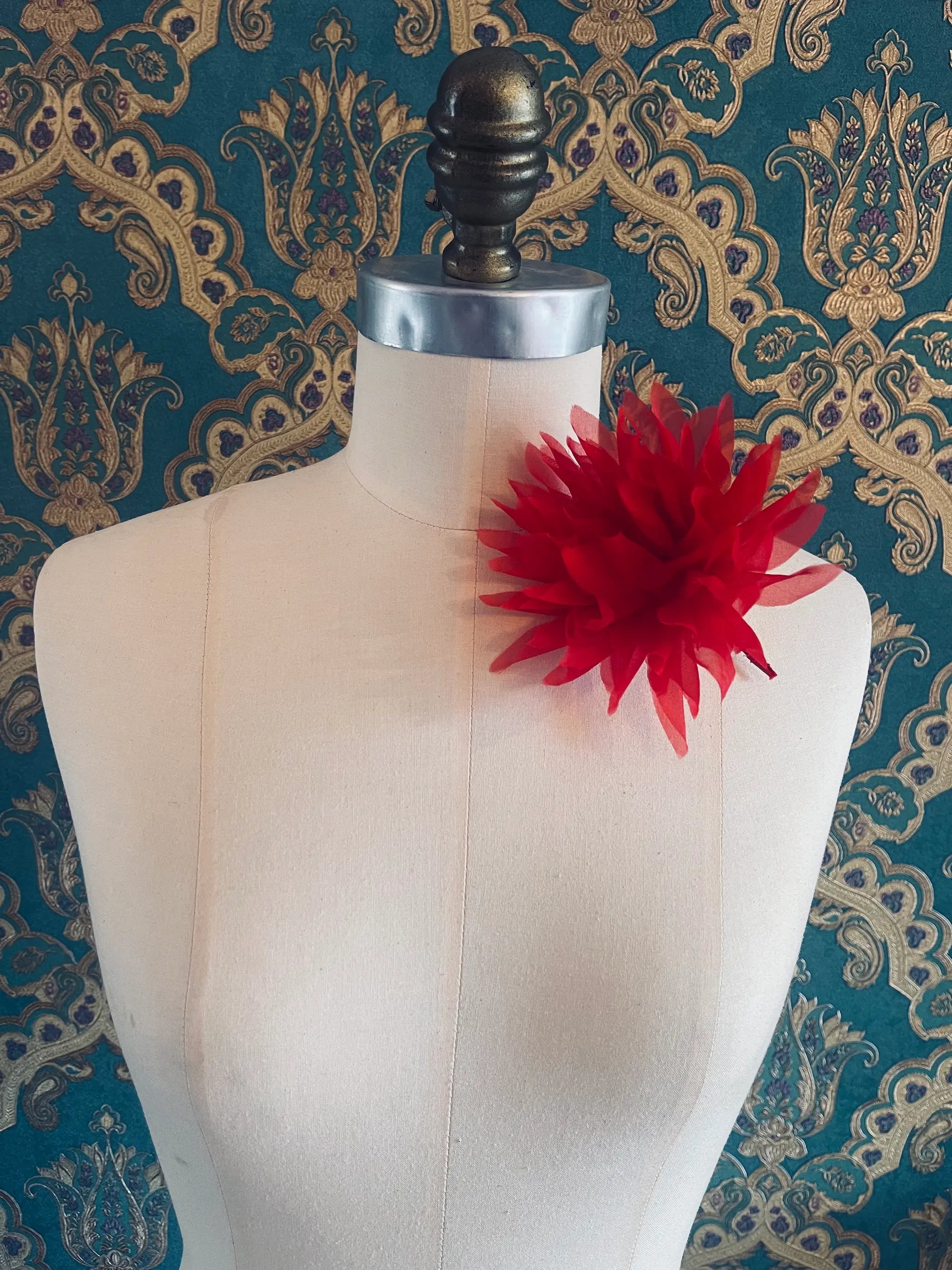 Broochella Dahlia  handmade fabric flower brooch in red. Styled on a mannequin. The background is green and gold damask wallpaper