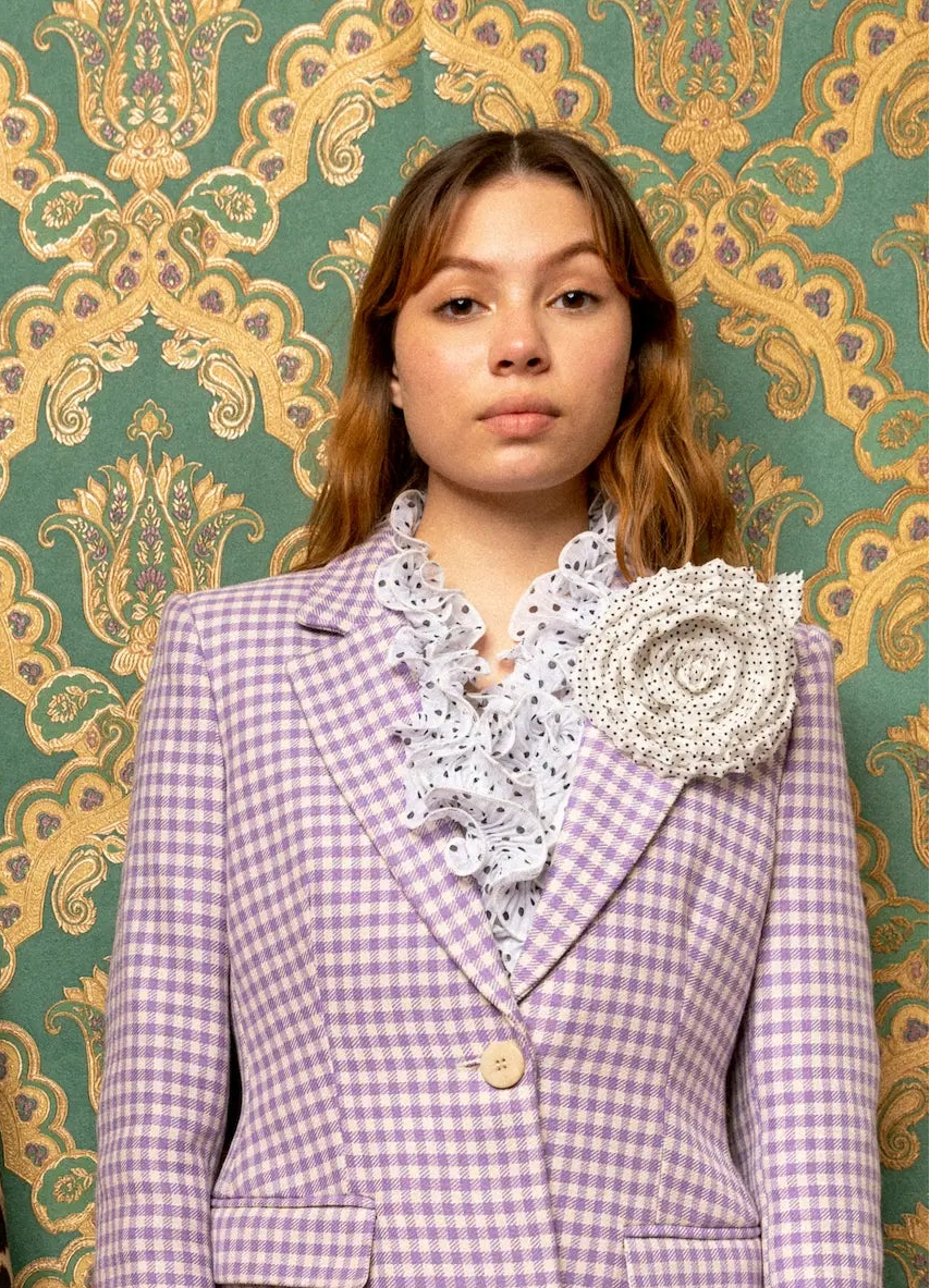 Two models standing side-by-side in front of a green and gold damask wallpaper background. The model on the right wears a purple checked jacket. She has the Broochella dalmata polka dot flower brooch pinned to her chest. 