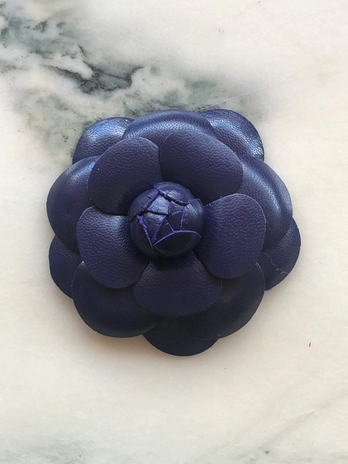A Broochella Camelia handmade dark purple leather flower-shaped accessory brooch,  resting on a white marble surface.