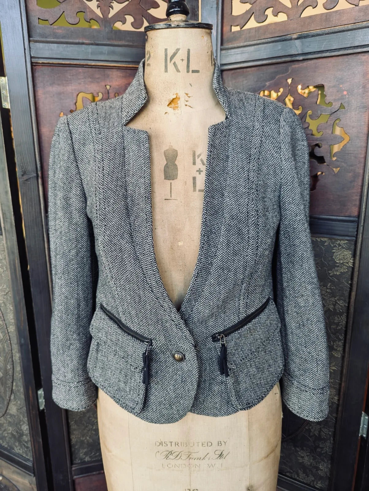 Broochella Day by Birger et Mikkelsen grey herringbone tweed jacket with black zip pockets. It is pictured on a mannequin. The background is an ornate dark wood room divider. 