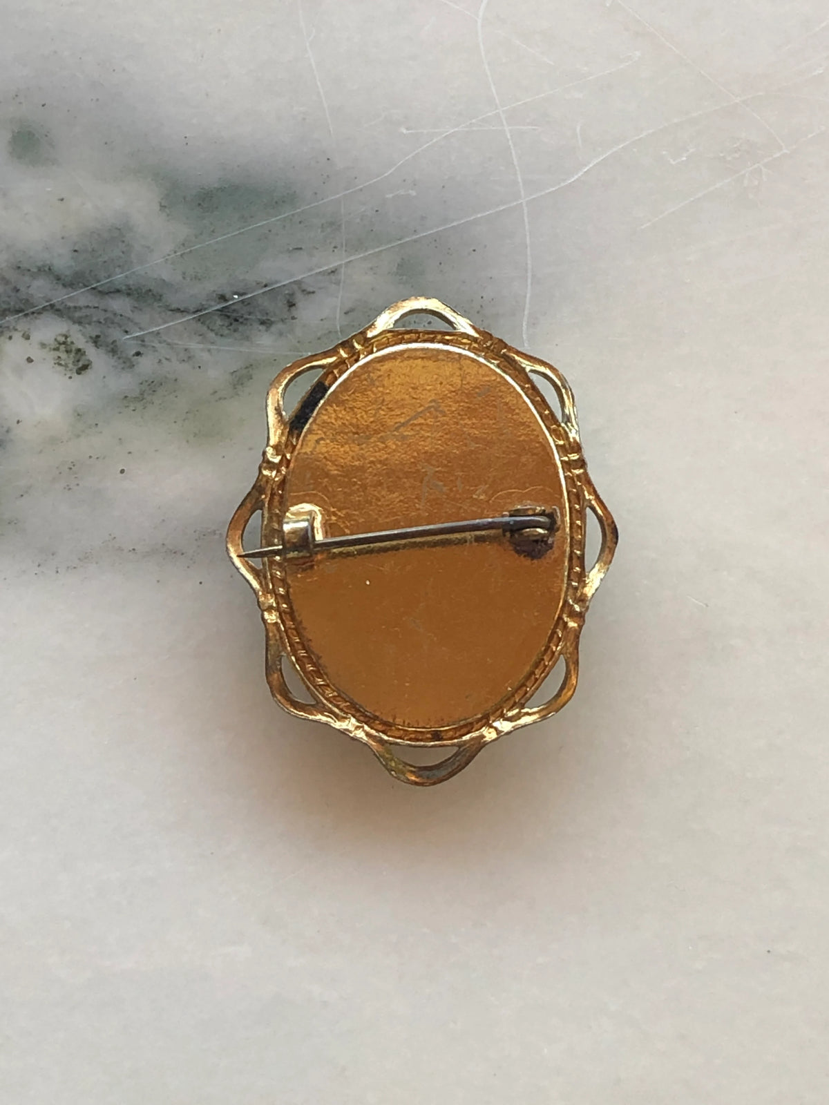 A Broochella Dea 4cm x 3cm vintage black and white cameo brooch featuring a carved white relief classical portrait of a noblewoman in profile with elaborate feathered and pearl upswept hair and a pearl necklace, set on a deep black oval ground within a gold-tone openwork filigree frame. The background is white marble