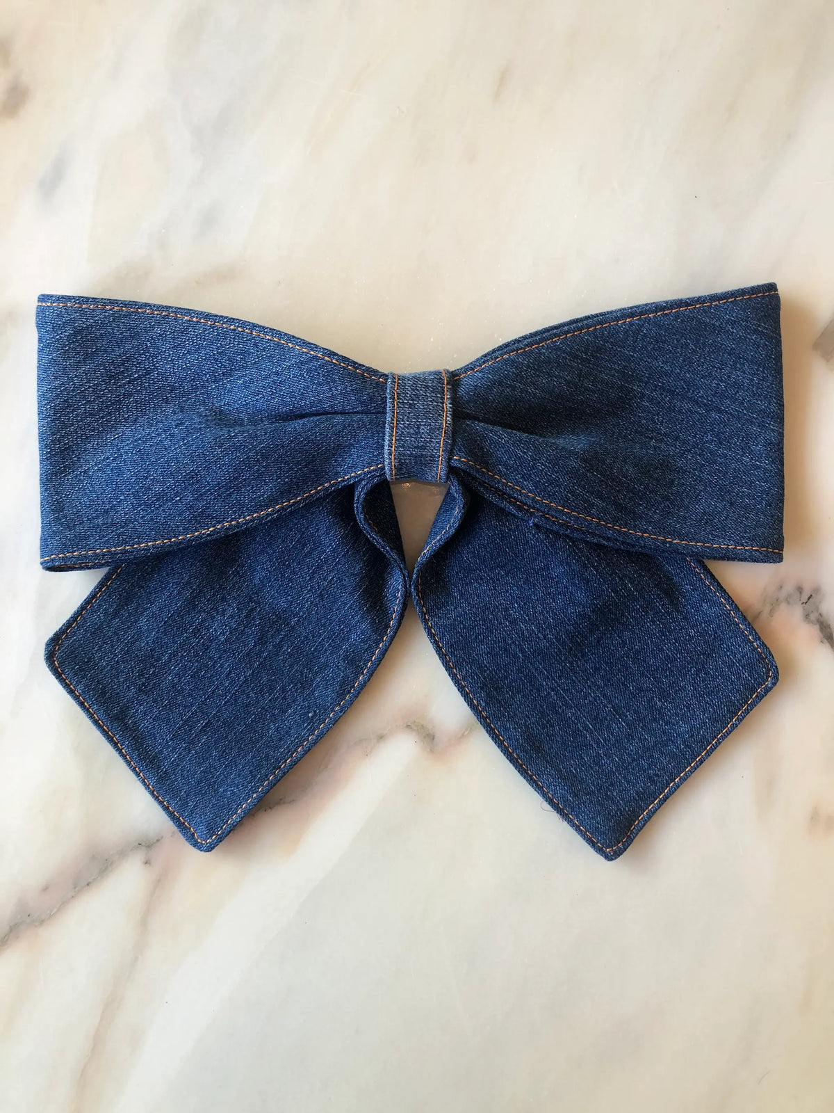 Broochella Denim Bow Brooch in dark blue denim with contrast tan stitching, featuring an oversized structured bow and long pointed tails, photographed on white marble.