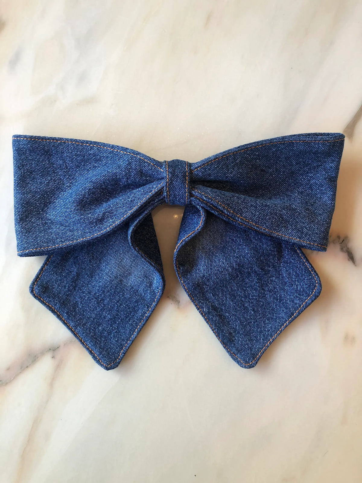 Broochella Denim Bow Brooch in dark blue denim with contrast tan stitching, featuring an oversized structured bow and long pointed tails, photographed on white marble.