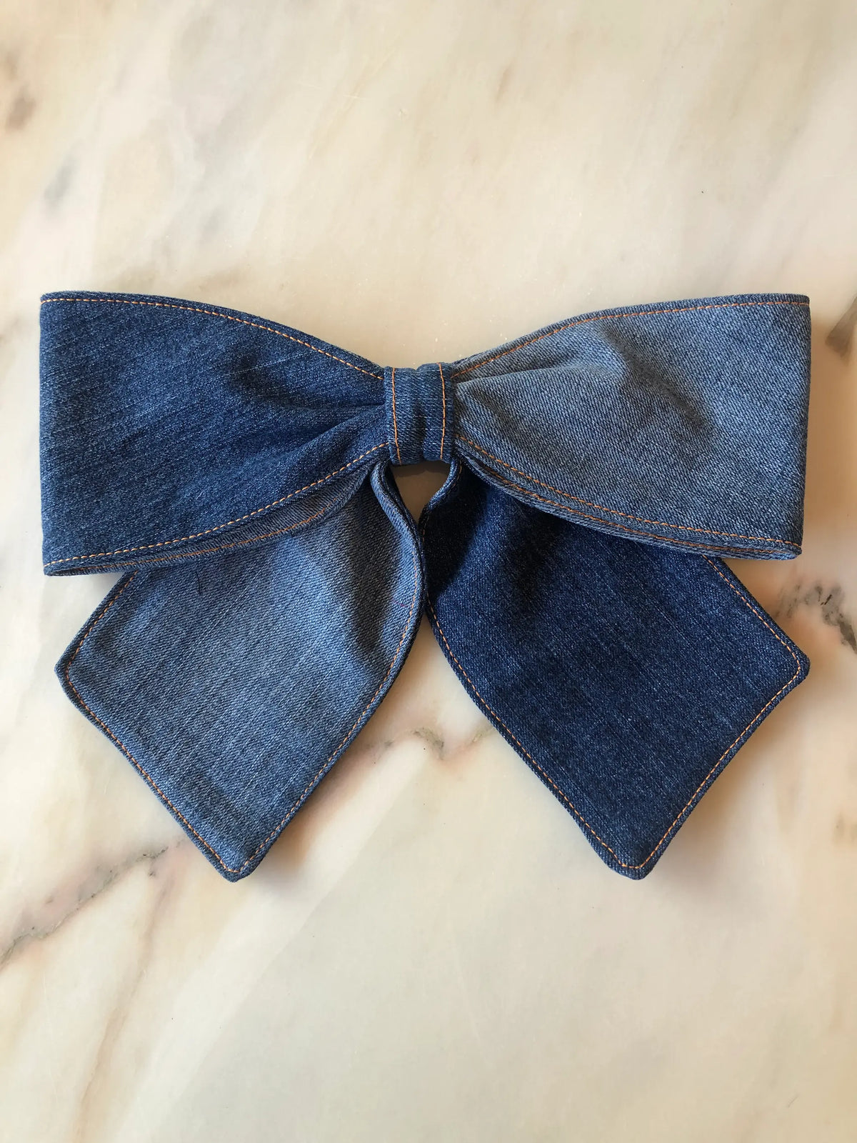 Broochella Denim Bow Brooch in dark blue denim with contrast tan stitching, featuring an oversized structured bow and long pointed tails, photographed on white marble.