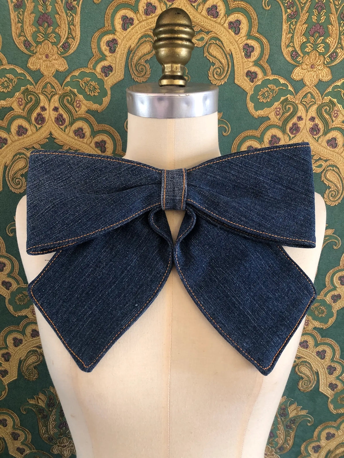 Broochella Large Denim Bow Brooch in dark blue denim with contrast stitching, displayed on a cream mannequin against ornate teal and gold wallpaper.