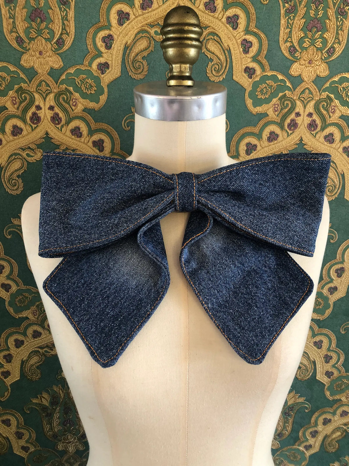 Broochella Large Denim Bow Brooch in dark blue denim with contrast stitching, displayed on a cream mannequin against ornate teal and gold wallpaper.