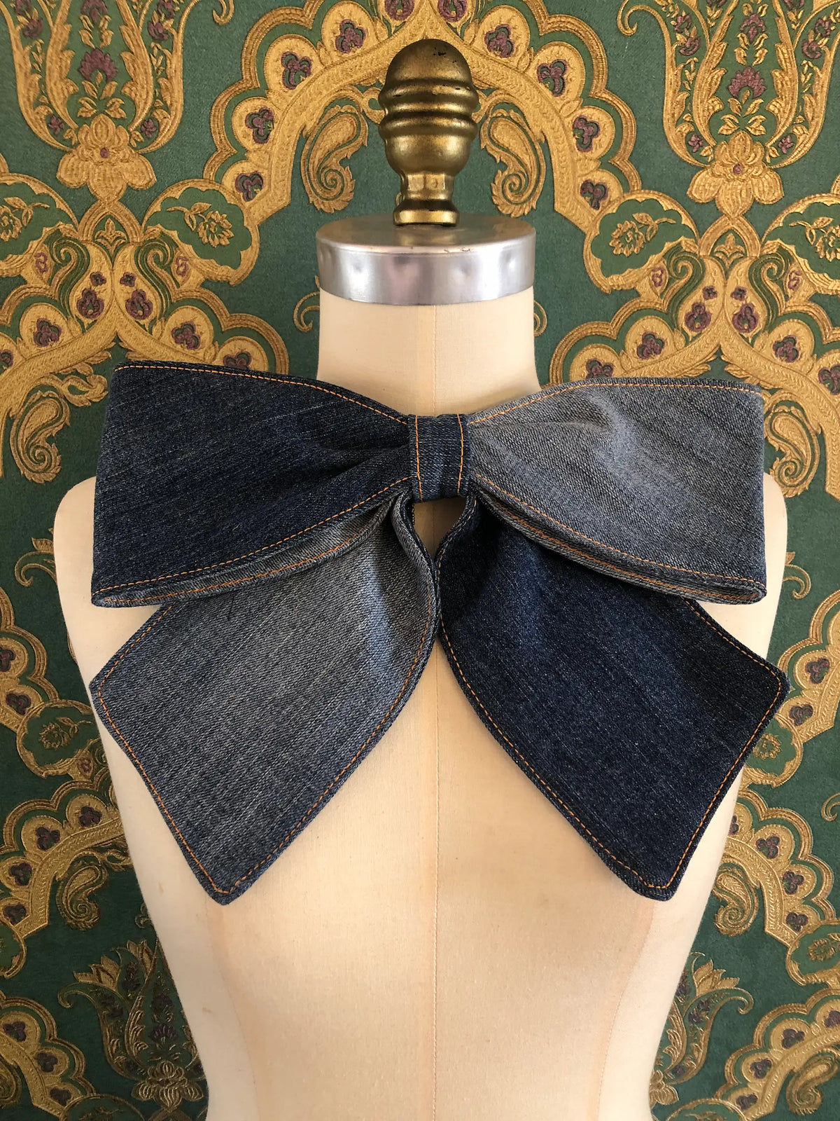 Broochella Large Denim Bow Brooch in dark blue denim with contrast stitching, displayed on a cream mannequin against ornate teal and gold wallpaper.