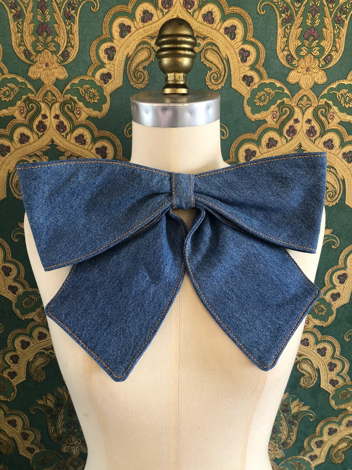 Broochella Large Denim Bow Brooch in dark blue denim with contrast stitching, displayed on a cream mannequin against ornate teal and gold wallpaper.