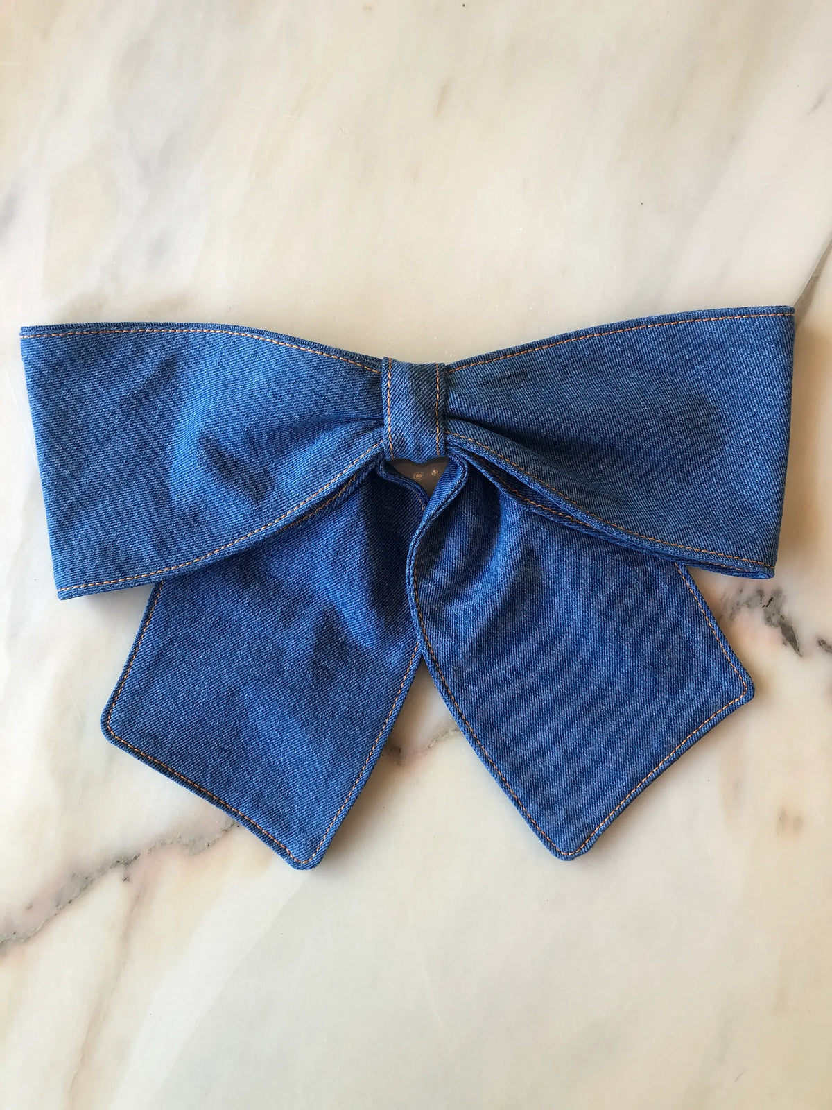 Broochella Denim Bow Brooch in mid-tone blue denim with contrast tan stitching, featuring an oversized structured bow and long pointed tails, photographed on white marble.