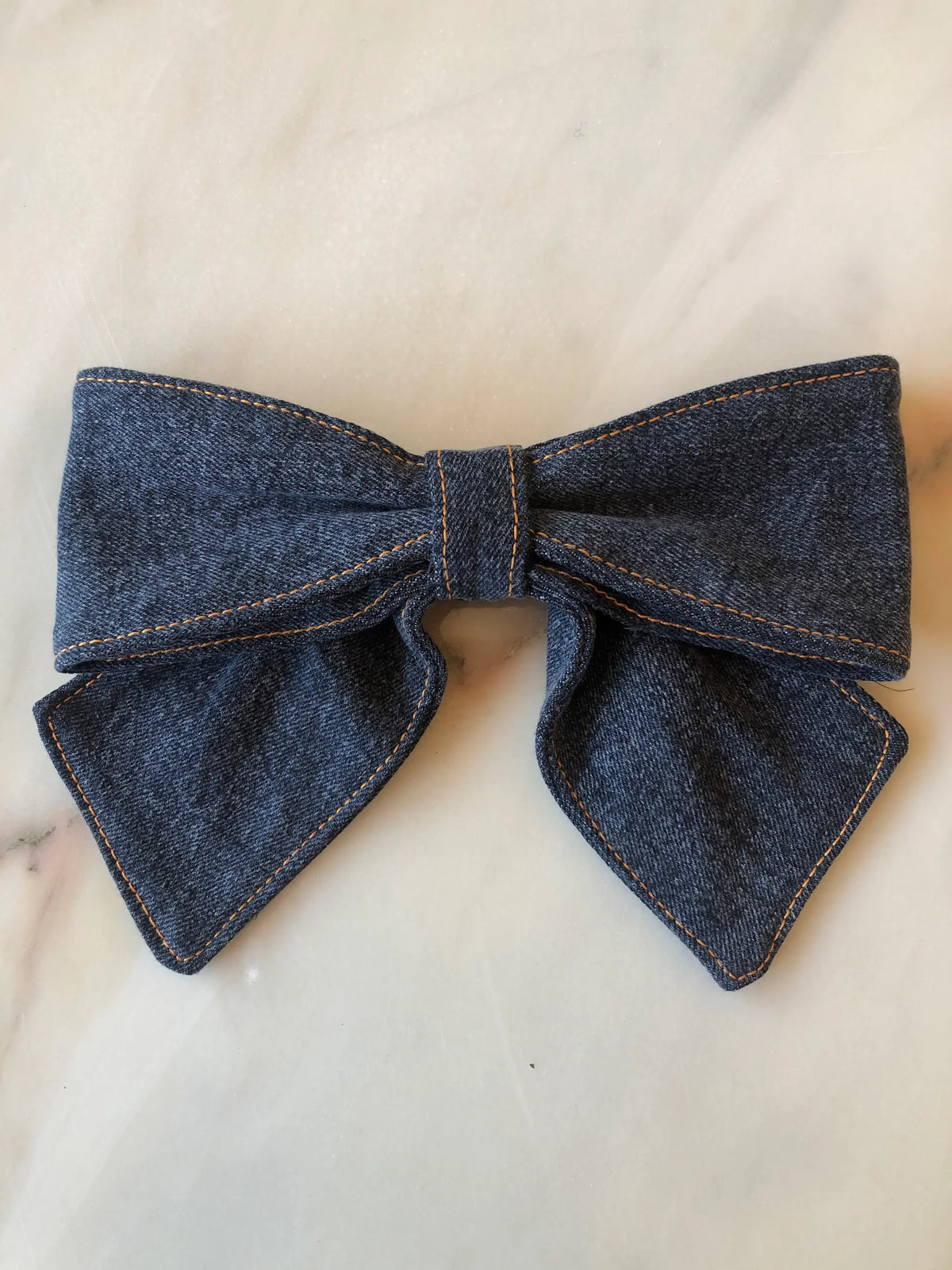 Broochella Medium Denim Bow Brooch in dark blue denim with contrast stitching, photographed on white marble.