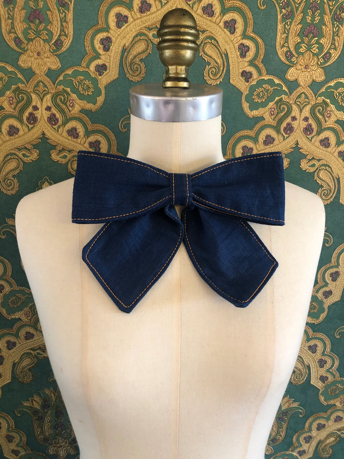 Broochella Medium Denim Bow Brooch in dark blue denim with contrast stitching, displayed on a cream mannequin against ornate teal and gold wallpaper.