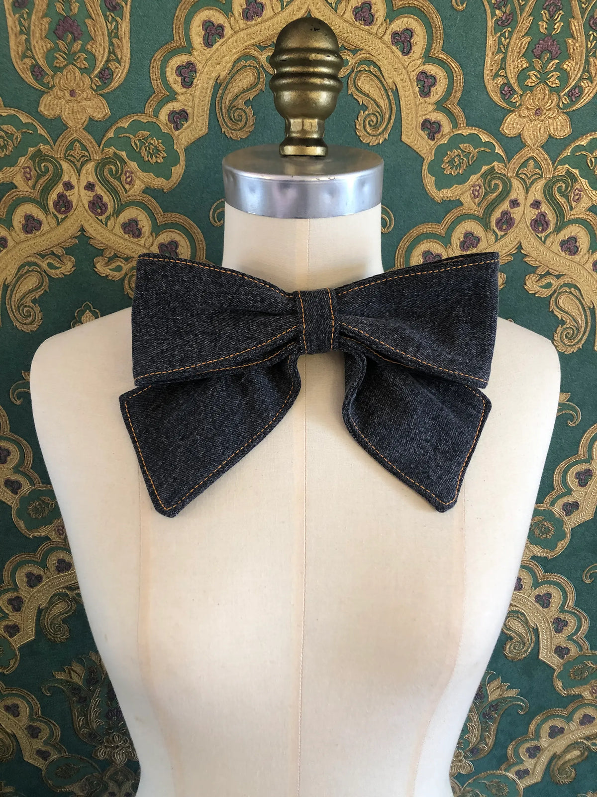 Broochella Medium Denim Bow Brooch in dark blue denim with contrast stitching, displayed on a cream mannequin against ornate teal and gold wallpaper.