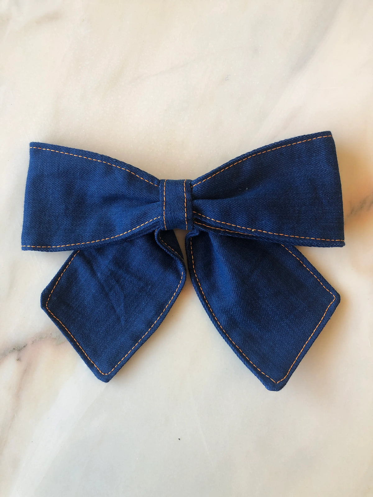 Broochella Medium Denim Bow Brooch in dark blue denim with contrast stitching, photographed on white marble.