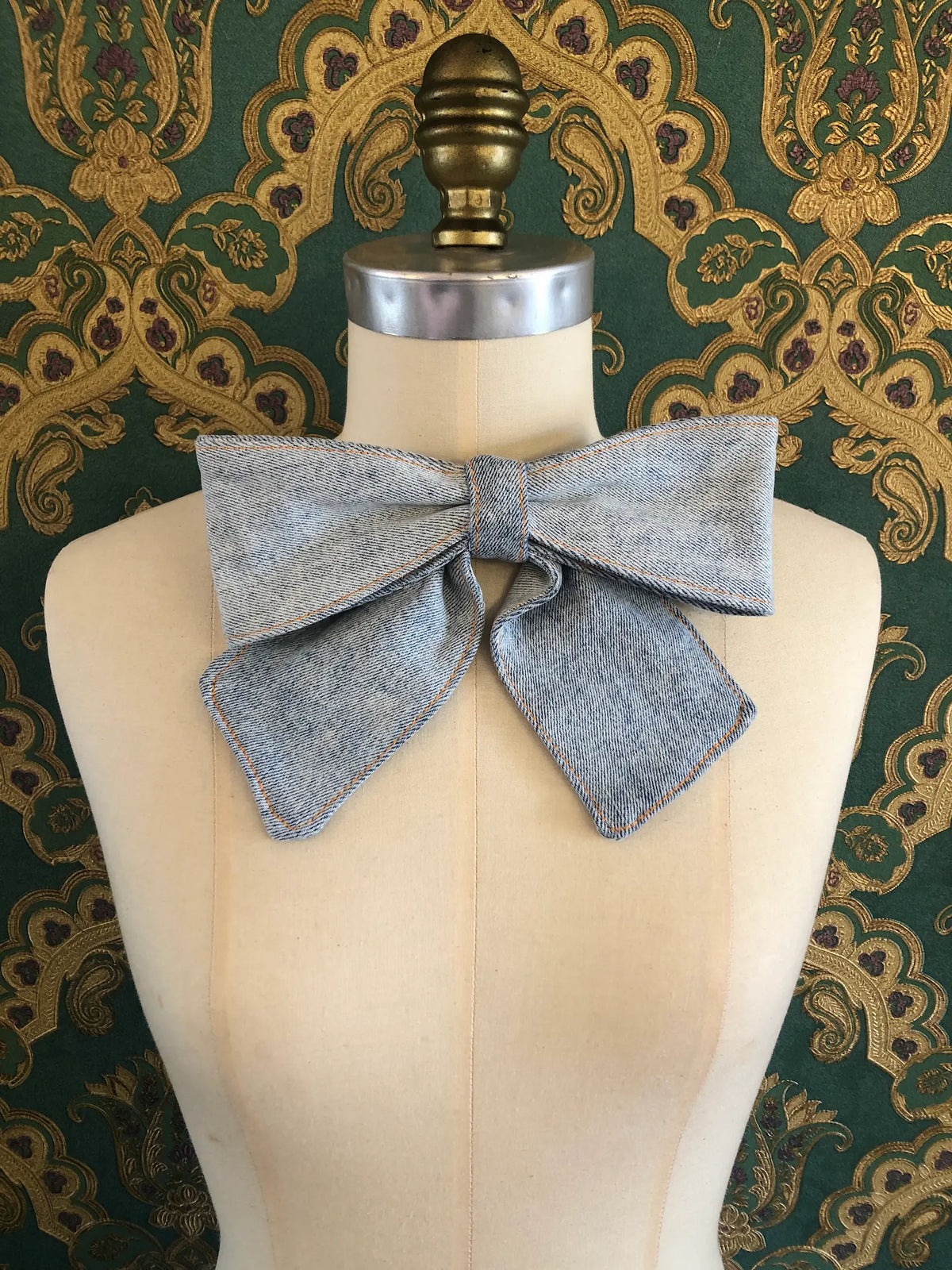 Broochella Medium Denim Bow Brooch in light blue denim with contrast stitching, displayed on a cream mannequin against ornate teal and gold wallpaper.