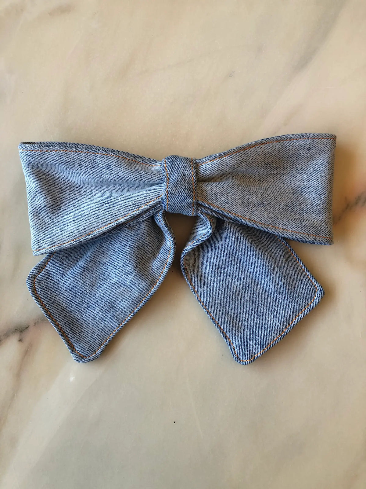 Broochella Medium Denim Bow Brooch in light blue denim with contrast stitching, photographed on white marble.