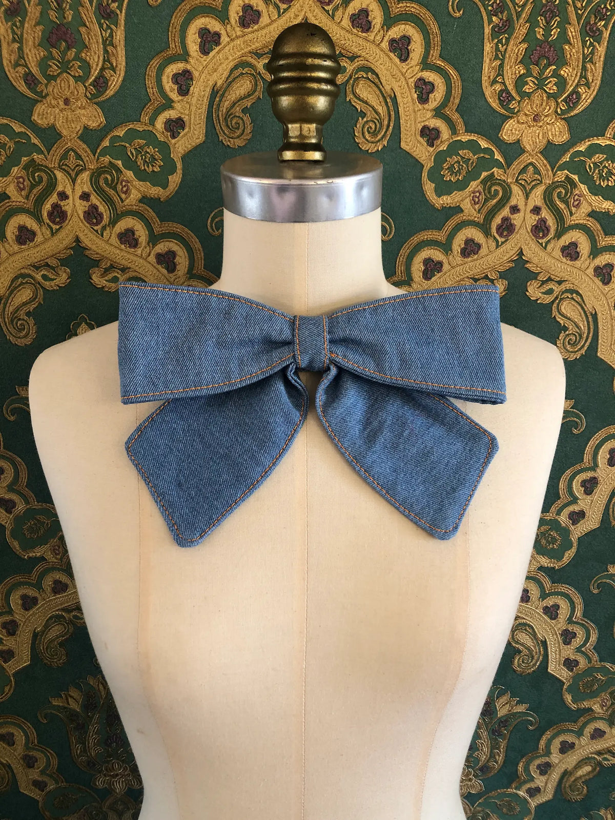 Broochella Medium Denim Bow Brooch in mid medium blue denim with contrast stitching, displayed on a cream mannequin against ornate teal and gold wallpaper.