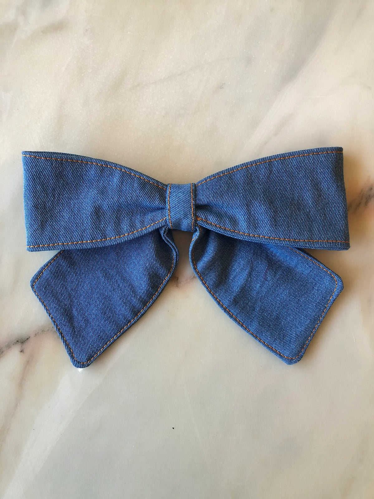 Broochella Medium Denim Bow Brooch in mid-blue denim with contrast stitching, photographed on white marble.