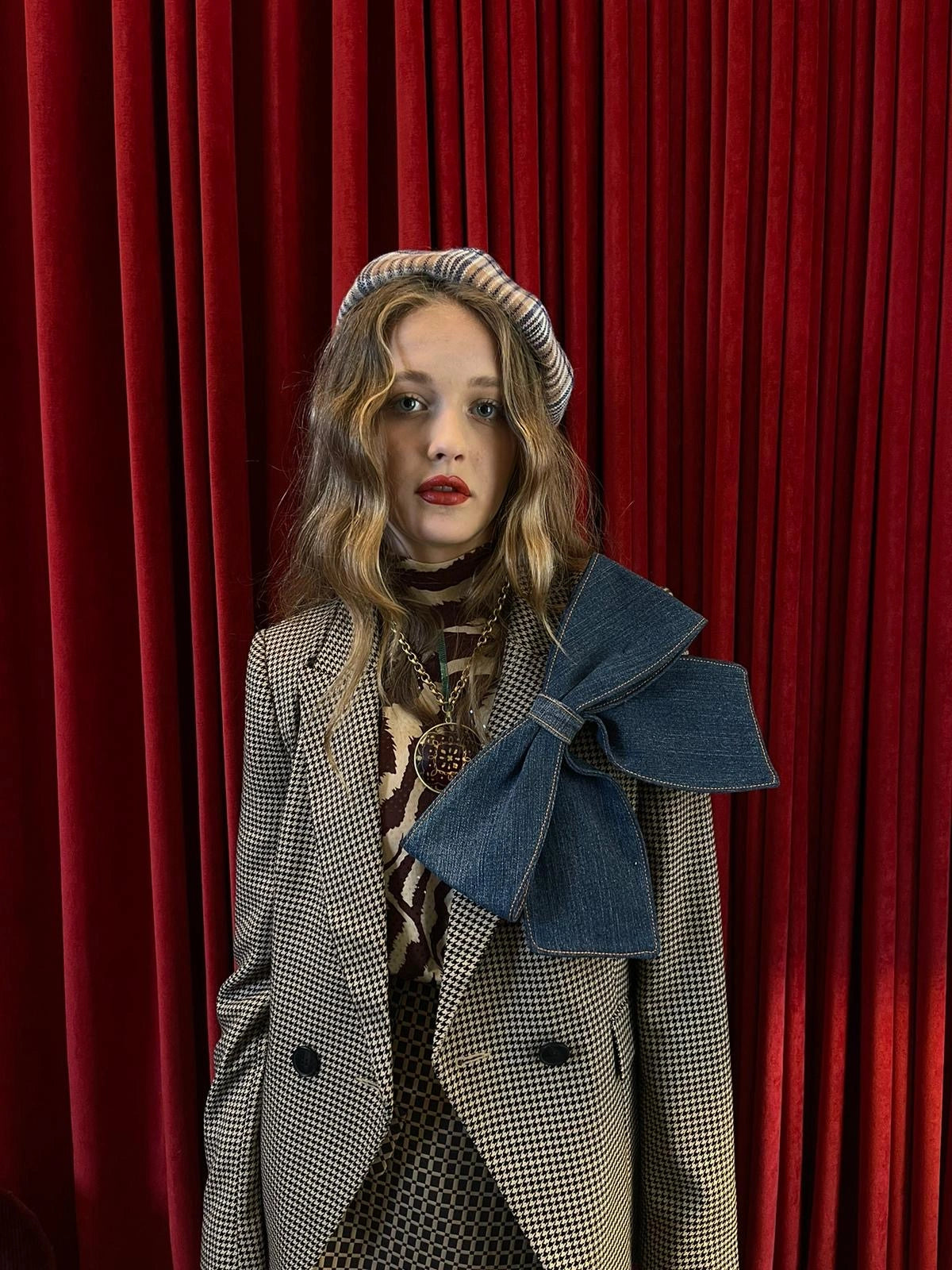 A model wearing a beige and black houndstooth blazer with a large blue Broochella denim bow on the lapel, a striped beret, and a patterned scarf, standing in front of a red velvet curtain.