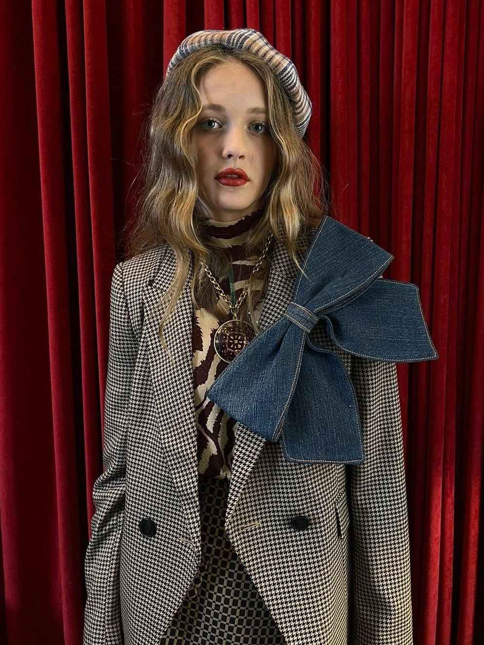 A model wearing a beige and black houndstooth blazer with a large blue Broochella denim bow on the lapel, a striped beret, and a patterned scarf, standing in front of a red velvet curtain.