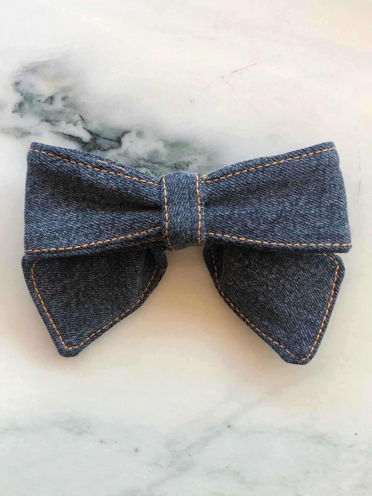 Broochella small black denim bow brooch pin with contrast tan stitching, photographed on a white marble background.