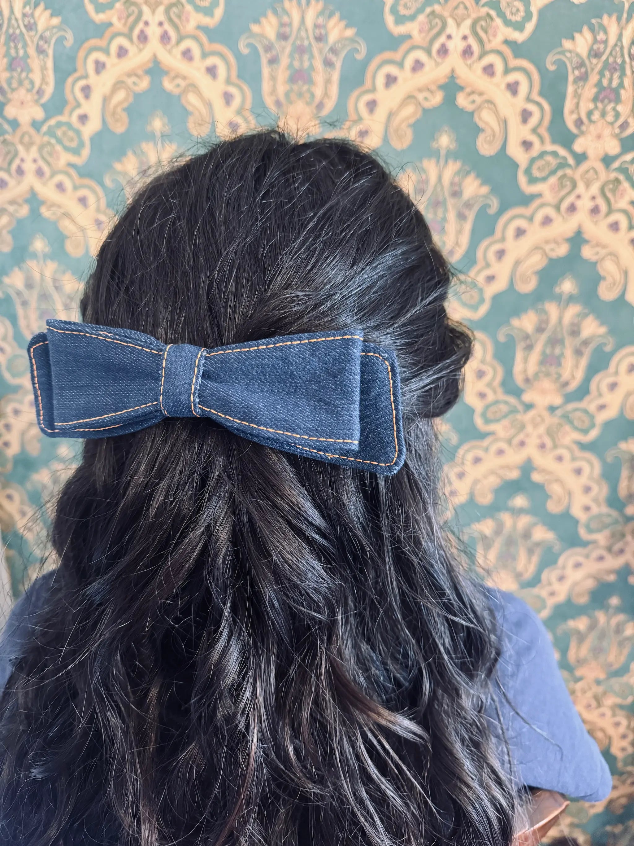 Broochella blue denim hair bow made from up cycled jeans. Pictured on a mannequin. The background is green and gold damask wallpaper.