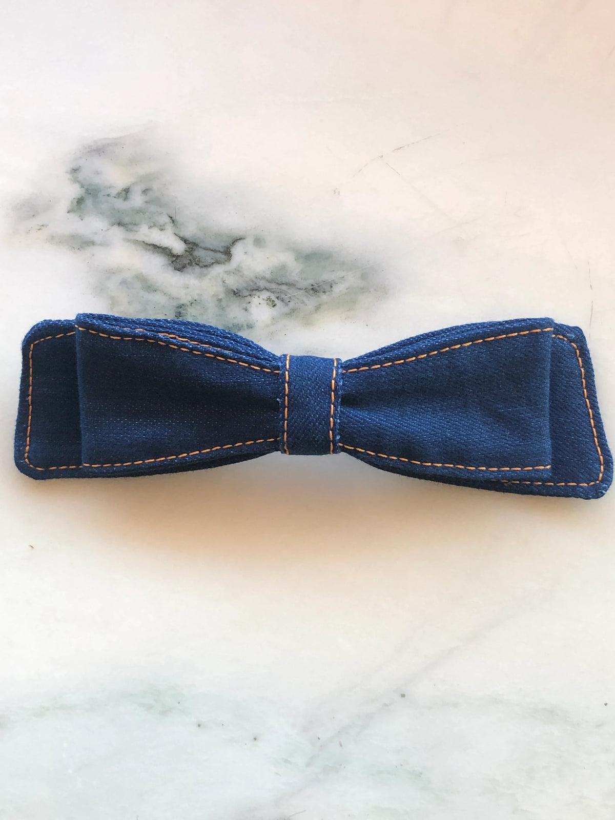 Broochella Denim Hair Bow in dark indigo denim with contrast tan stitching and structured double bow design, photographed on white marble.