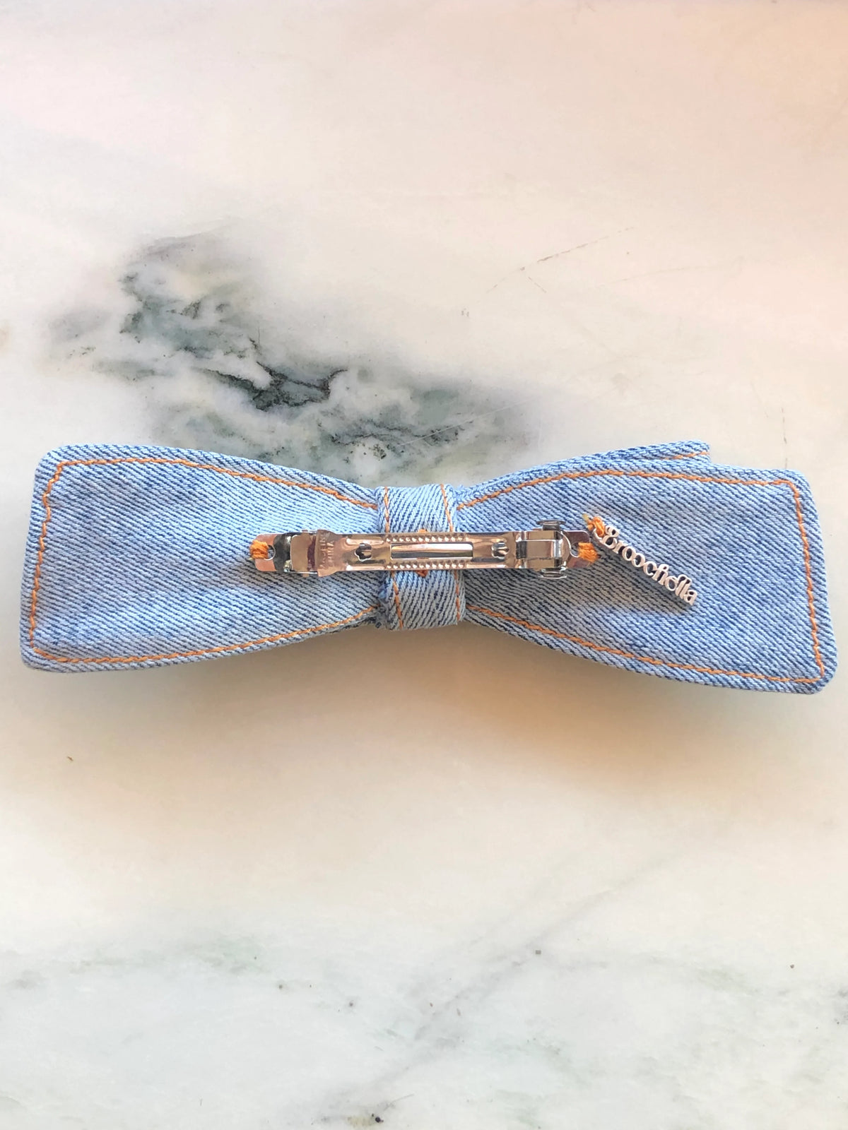 The back view of Broochella handmade Denim Hair Bow in light blue denim with contrast tan stitching, photographed on white marble.