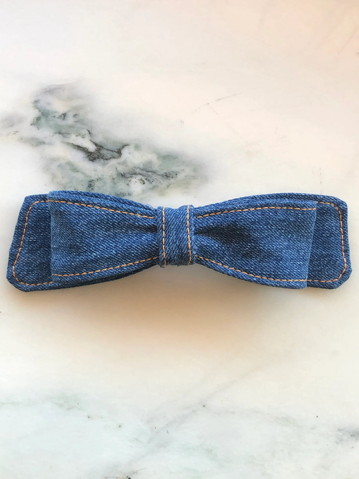 Broochella Denim Hair Bow in mid-blue denim with contrast tan stitching and structured double bow design, photographed on white marble.