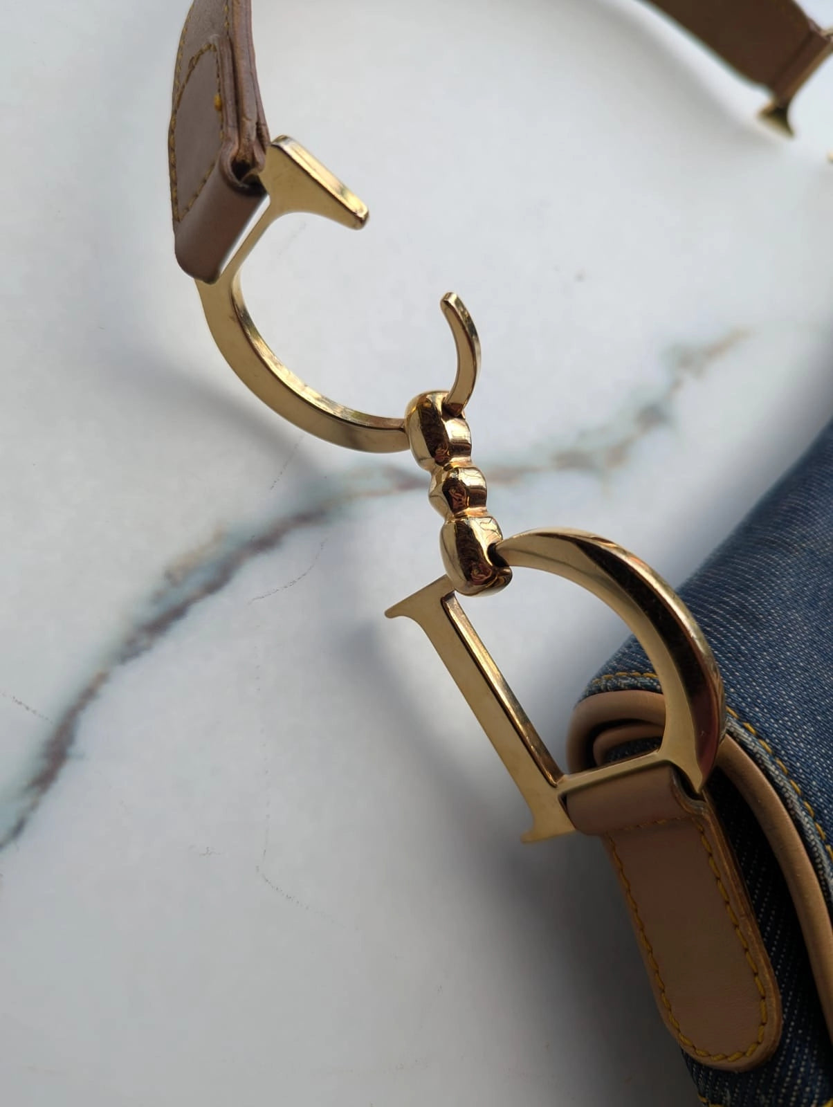 Broochella Dior Authentic Vintage designer denim saddlebag with gold metal logo fixings and brown leather straps. It is pictured on a white marble surface. 