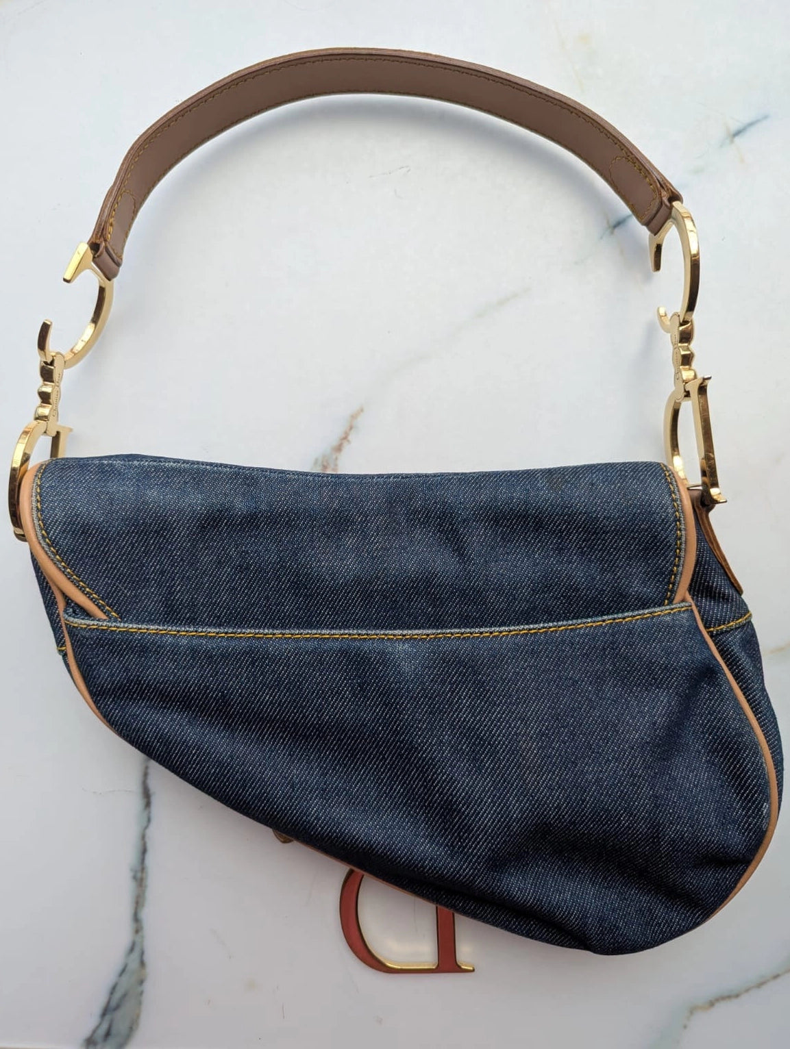 Broochella Dior Authentic Vintage designer denim saddlebag with gold metal logo fixings and brown leather straps. It is pictured on a white marble surface. 