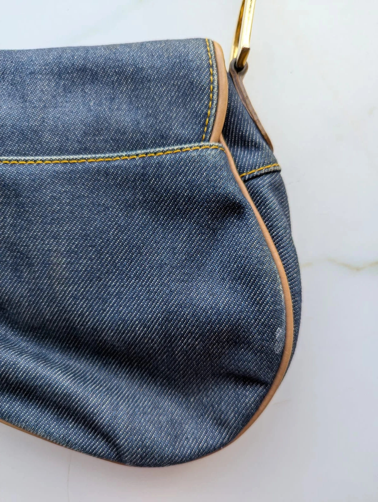 Broochella Dior Authentic Vintage designer denim saddlebag with gold metal logo fixings and brown leather straps. It is pictured on a white marble surface. 