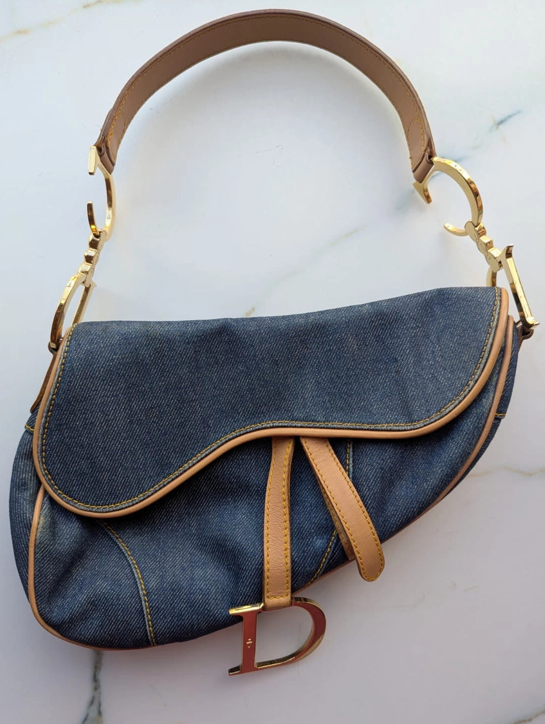 Broochella Dior Authentic Vintage designer denim saddlebag with gold metal logo fixings and brown leather straps. It is pictured on a white marble surface. 