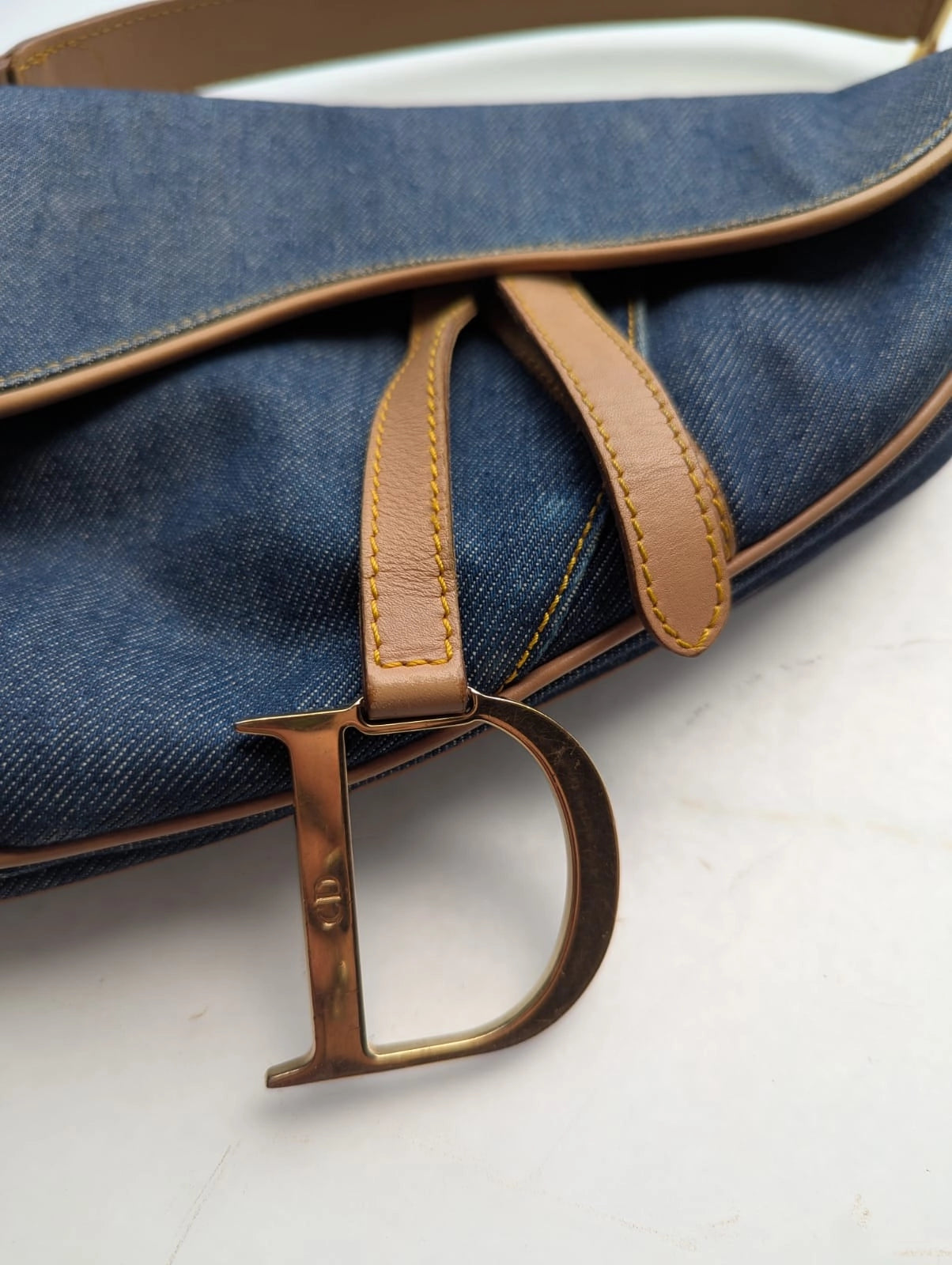 Broochella Dior Authentic Vintage designer denim saddlebag with gold metal logo fixings and brown leather straps. It is pictured on a white marble surface. 