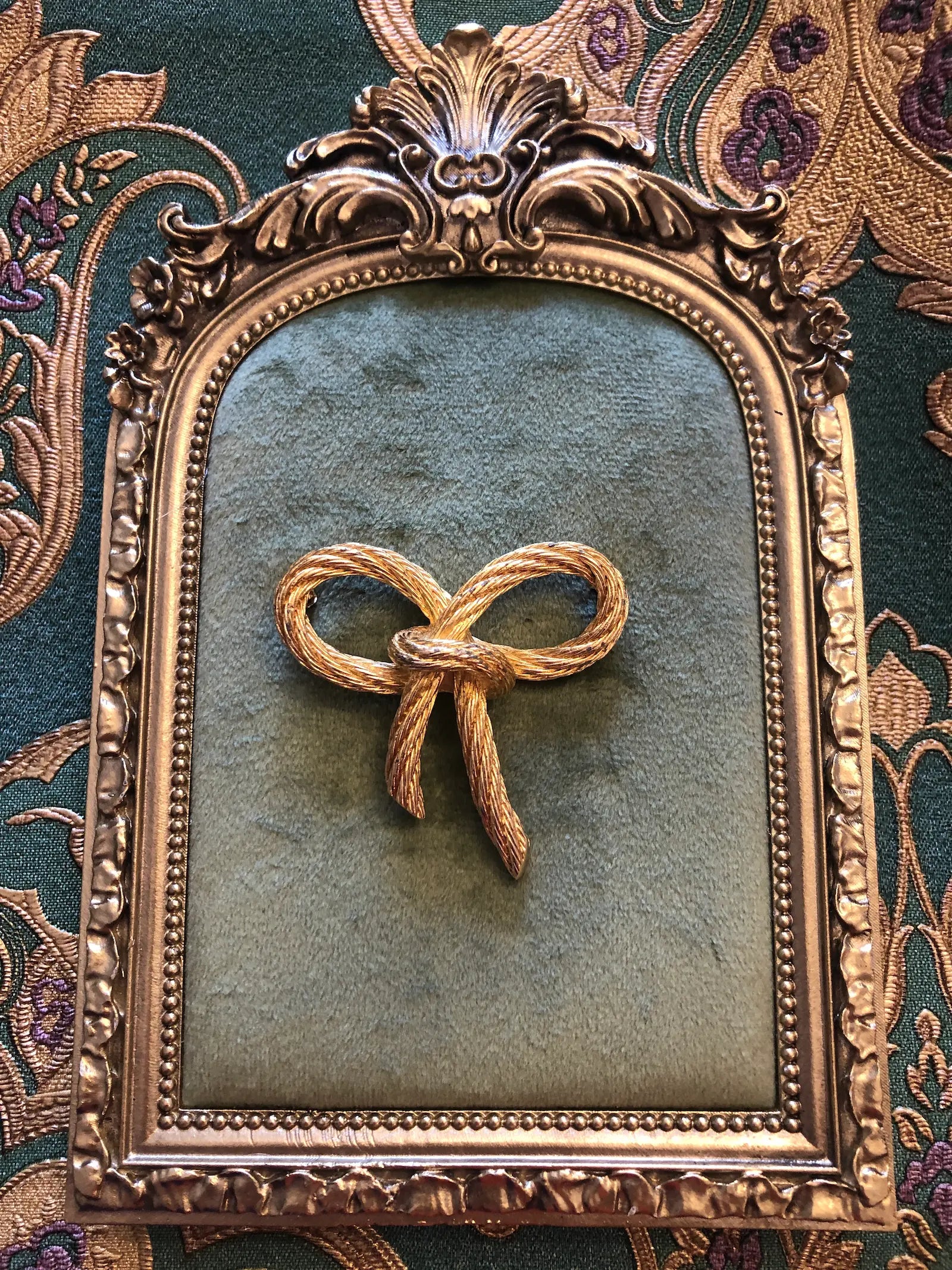 Broochella vintage authentic dior costume bow brooch in gold twisted metal rope. Pictured on a green velvet tray with a gold frame. The background is green and gold damask wallpaper. 